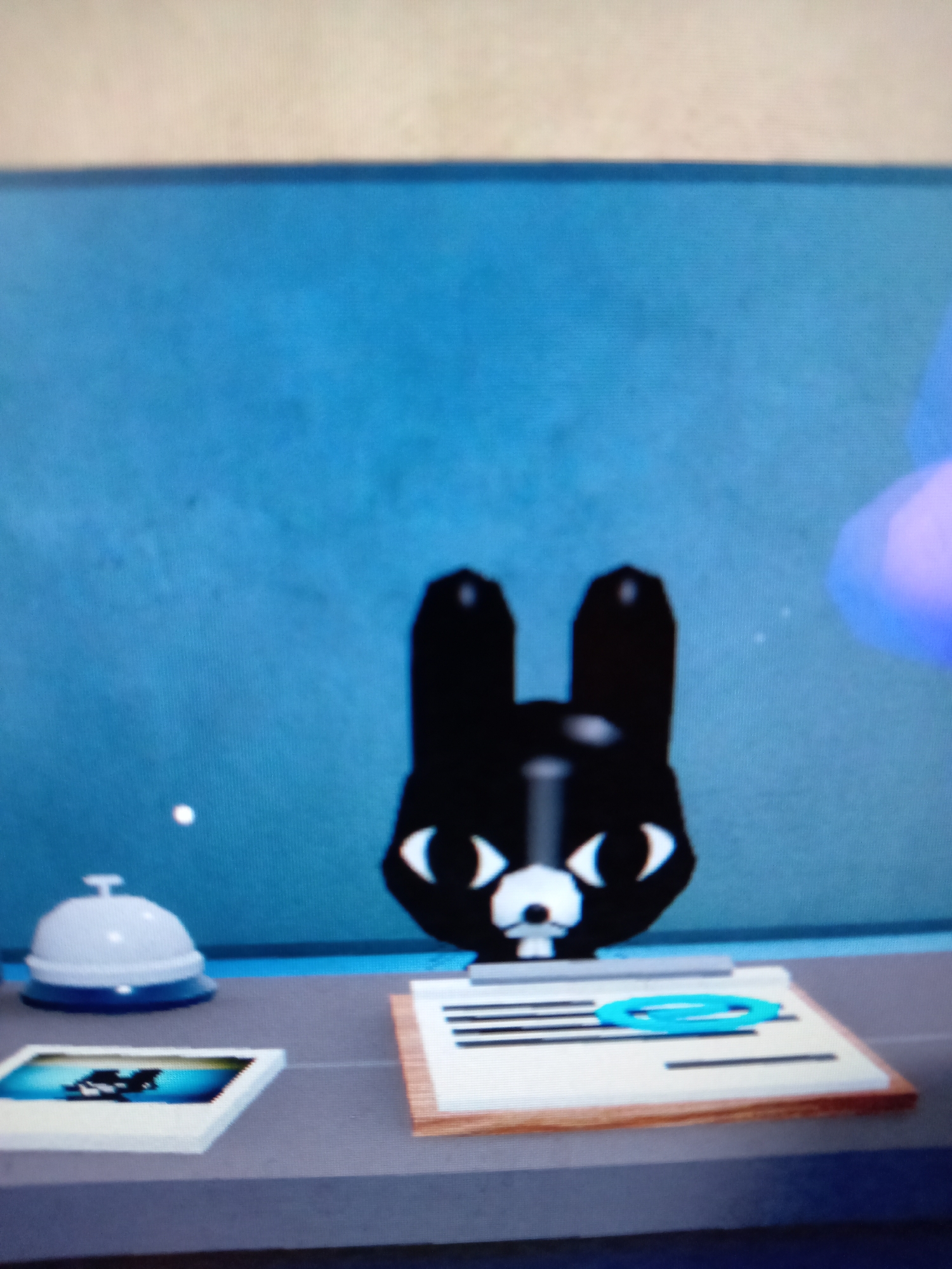 This is a close-up, slightly blurry, eye-level photograph of a cartoon black cat character on a counter. The cat has large white eyes with black pupils, a white muzzle with a small black nose and mouth, and two tall black ears. The background is a solid blue, possibly a wall.
On the counter in front of the cat are three objects. To the left is a white desk bell with a silver handle. Next to the bell is a framed picture, possibly a photo or a print, with a black and white image of what appears to be a cat or a similar creature. To the right of the picture is a clipboard with a wooden border and white paper, on which several black lines are drawn. A bright blue paperclip is resting on the clipboard. To the right of the clipboard, a blurred purple and blue object is visible, which might be a hand or another digital element.
The image does not provide any information about the location in Rudny, Kazakhstan.