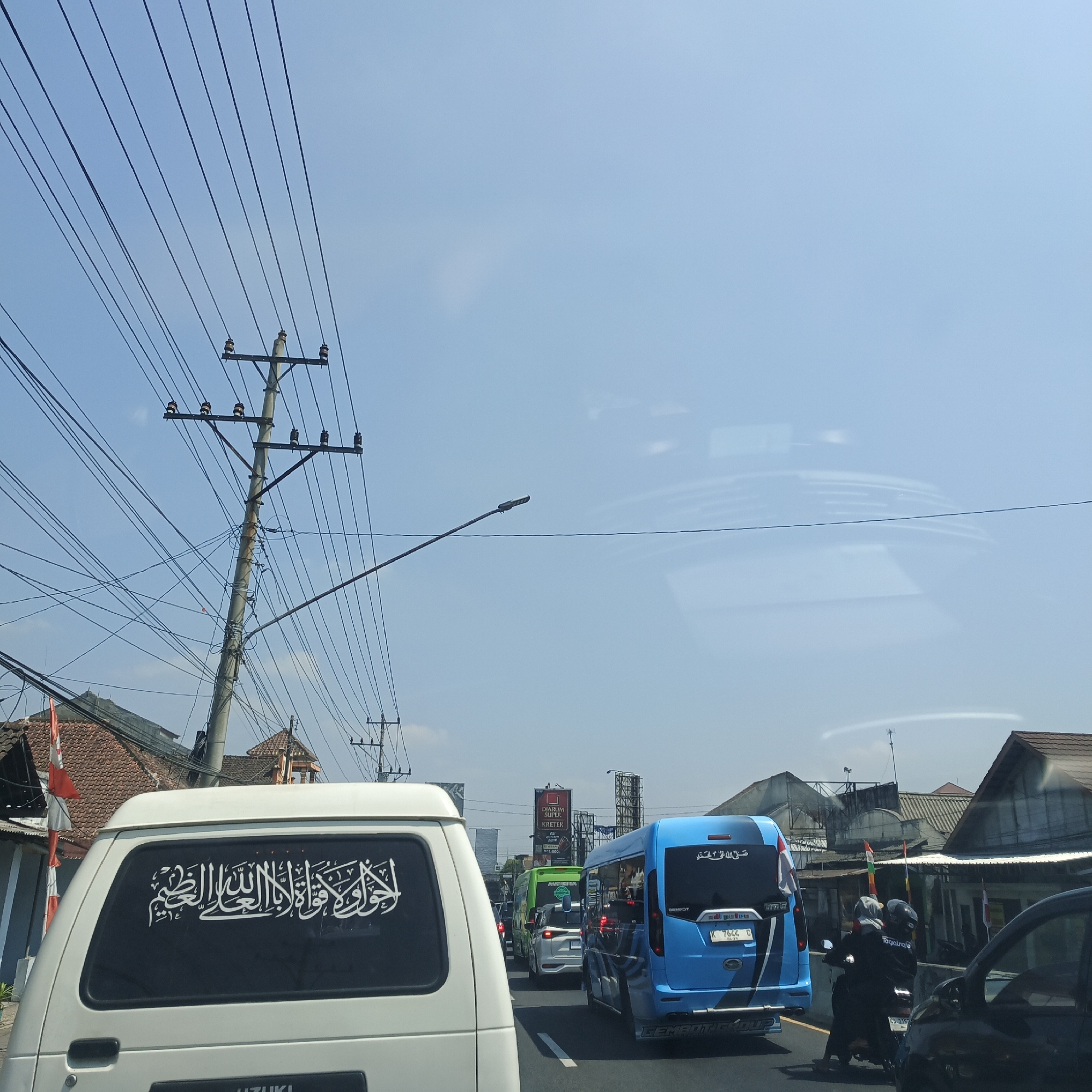 In Secang, Indonesia, a traffic jam is depicted on a sunny day. The foreground is dominated by the rear of a white van with a black window featuring white Arabic calligraphy. To the right, a bright blue bus is visible with similar Arabic lettering on its rear window, and a motorcycle with two occupants, one wearing a grey helmet and the other a dark helmet, is positioned behind it. Further down the road, a green bus and a white car are also caught in the traffic. The scene is characterized by an abundance of overhead power lines strung from a tall, concrete utility pole on the left, with additional poles and lines stretching across the frame. Buildings with terracotta roofs line the sides of the road, and billboards are visible in the background. The sky is clear and blue with some faint reflections on a glass surface, possibly from the camera's perspective.