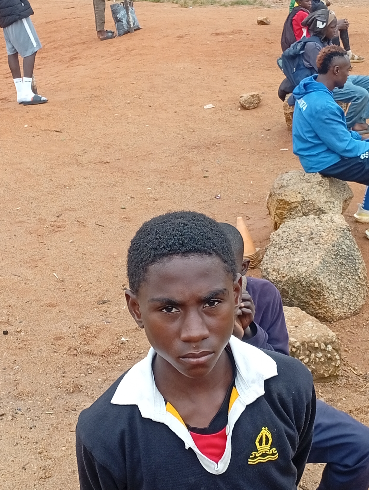 A group of boys and young men are gathered outdoors on a dirt ground. The foreground features a young boy with dark, curly hair, wearing a dark sweater with a white collar and a yellow and red trim. He has a golden crest on his sweater. Behind him, another boy in a purple long-sleeved shirt peeks out. To the right, a young man with styled hair wears a bright blue hooded jacket with white text on the sleeve. Other individuals are visible in the background, some sitting on rocks. One person on the left is wearing grey shorts and white socks with sandals. The environment appears to be a dry, reddish-brown earth, with scattered rocks and some vegetation. The scene suggests a casual gathering, possibly a school or community event, taking place in a rural or semi-urban area of Jos, Nigeria.