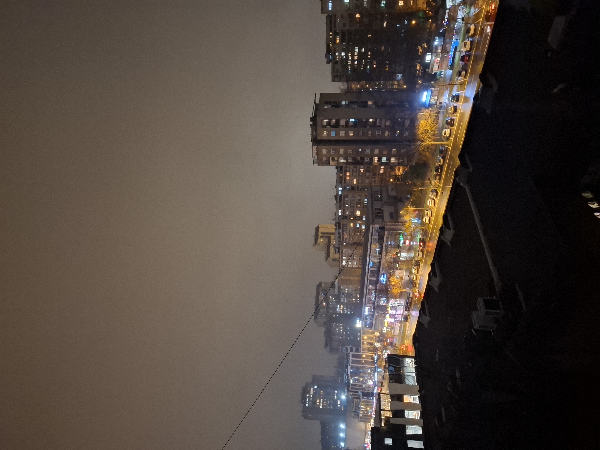 This is a nighttime photograph taken in Novi Sad, Serbia. It depicts a view of the city's skyline, with a focus on a street lined with illuminated buildings. The sky is overcast, casting a dim light over the scene. There are multiple residential and commercial buildings. The street appears to be a major thoroughfare with traffic.