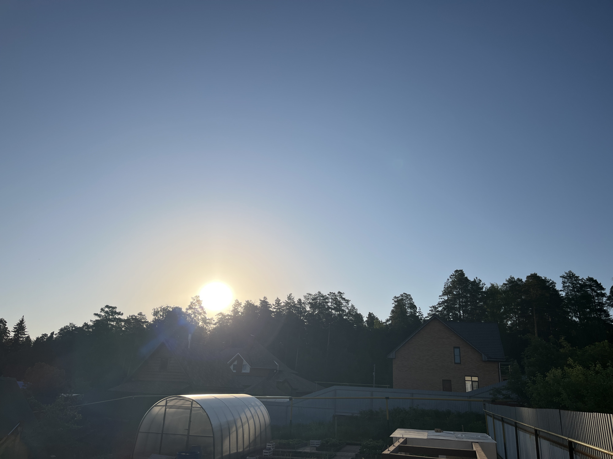 The image captures an outdoor scene in Chaikovsky, Russia, at what appears to be either sunrise or sunset, based on the low position of the sun. The sky is mostly clear blue. The sun is partially obscured by a dense line of trees. Below the treeline are several structures: a greenhouse with a translucent arched roof, a wooden house, and a two-story brick house with a tiled roof. A metal fence runs across the lower portion of the image. The foreground contains some building materials or construction debris, suggesting ongoing work or recent activity. The scene conveys a sense of rural or suburban living adjacent to a forested area.