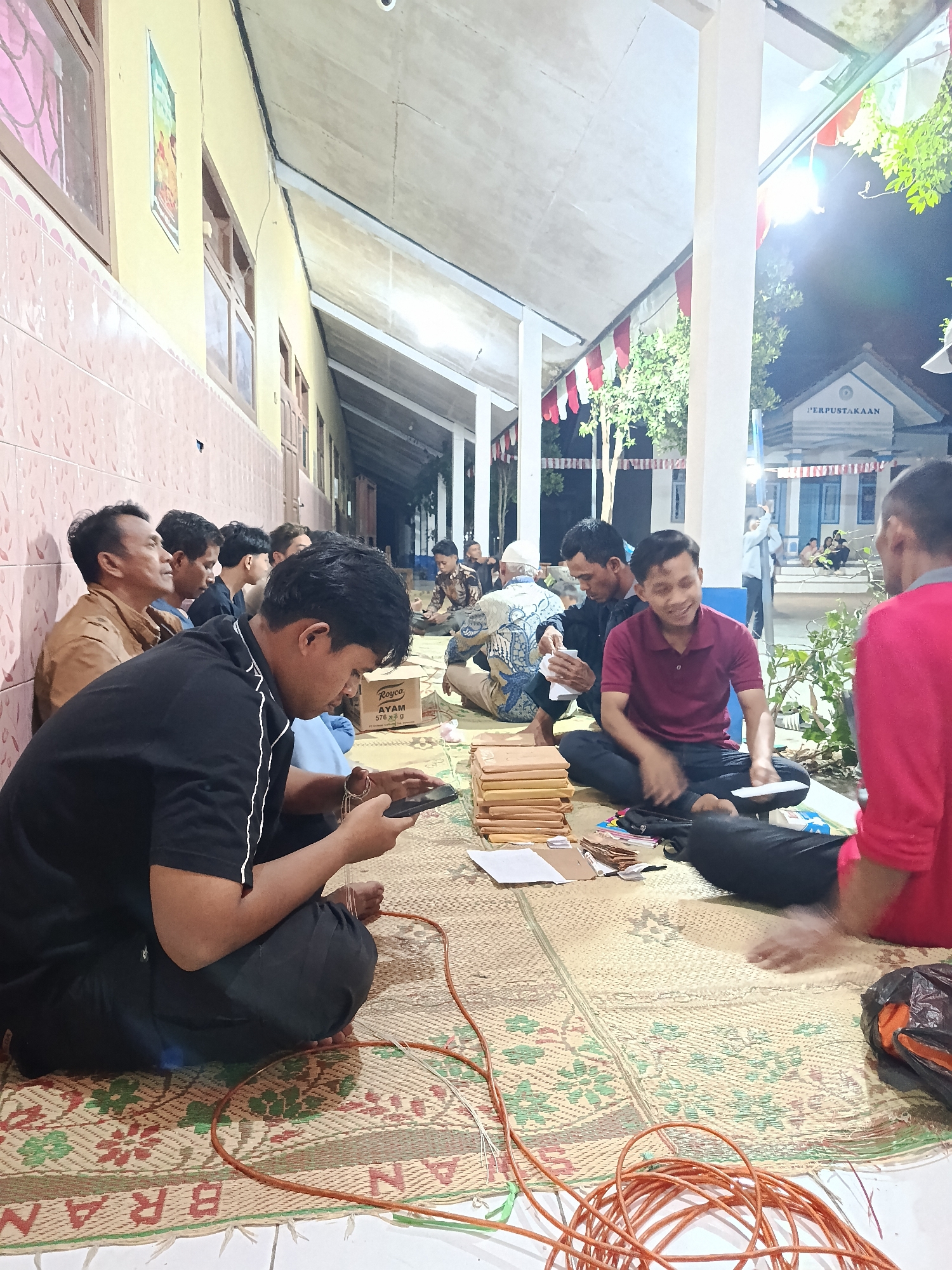 In Pocol, Indonesia, a group of men are gathered on woven mats in what appears to be the outdoor hallway of a building, possibly a library as indicated by a sign reading "PERPUSTAKAAN" on the structure in the background. The scene is lit by artificial lights and the ambient night sky, with red and white Indonesian flags strung overhead, suggesting a festive or commemorative occasion. Some men are seated on the mats, engaged in conversation or activity, while others are observing. One man in the foreground is focused on his mobile phone. Stacks of documents and various small objects are scattered on the mats, indicating some form of organized activity or gathering. The architectural style of the building, with its tiled facade and open-air corridor, is typical of Indonesian public buildings.