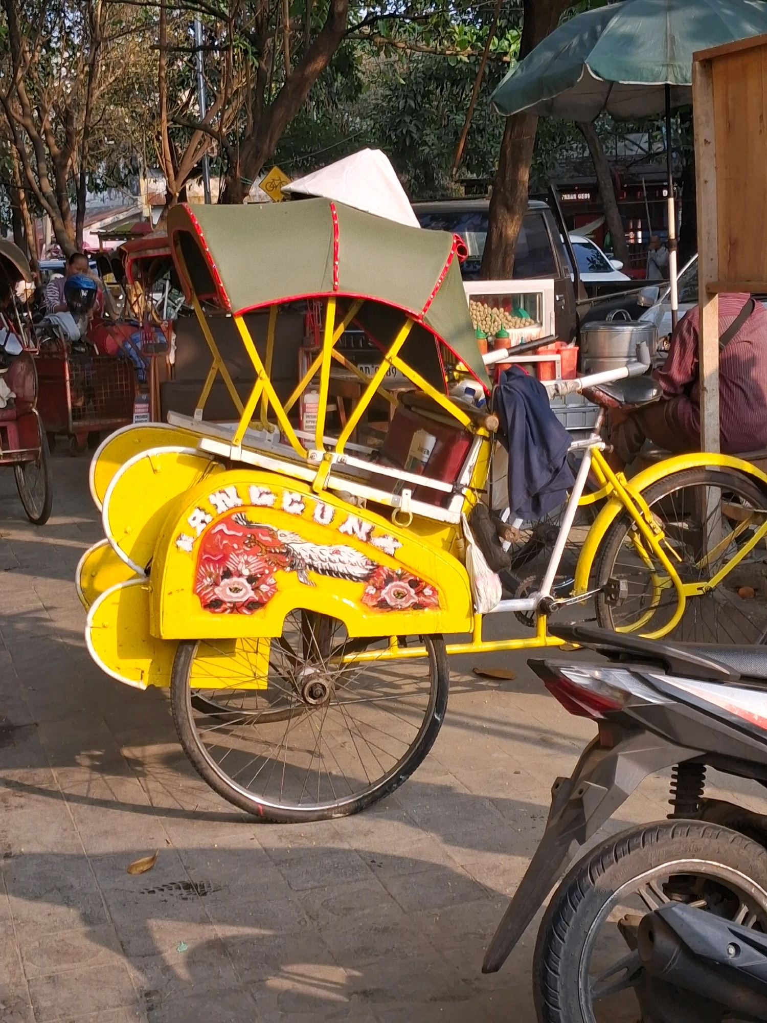 The image depicts a yellow pedicab, known locally as a "becak," parked on a paved street in Surakarta, Indonesia. The becak features a green canopy with red trim, supported by a yellow frame. A decorative image of a dragon is painted on its side. A person is seated on the becak, wearing a blue helmet. In the background, trees provide shade, and other vehicles, including a car and a motorcycle, are visible. The setting appears to be a street scene in a bustling city, possibly a market area given the presence of goods displayed in a glass case on a cart. The lighting suggests it is daytime, with shadows cast on the ground.