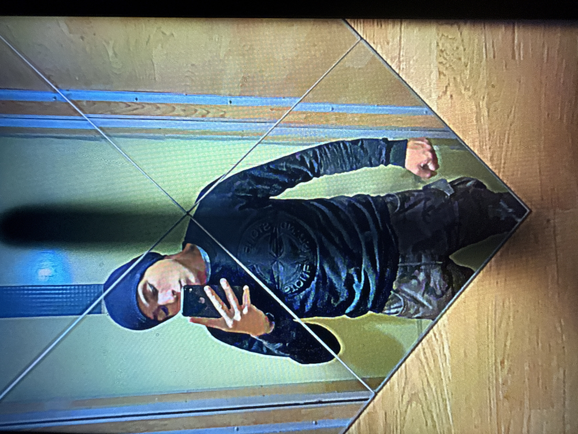A young man wearing a dark blue beanie and a dark long-sleeved shirt with a circular logo, and camouflage-patterned cargo pants, is taking a mirror selfie. He is holding a smartphone in his right hand, angled to capture his reflection. The background appears to be an elevator interior, with wooden paneling on the walls and a metal strip running diagonally across the mirror. The lighting is dim, casting shadows. The scene is a personal moment captured in a mundane urban setting.