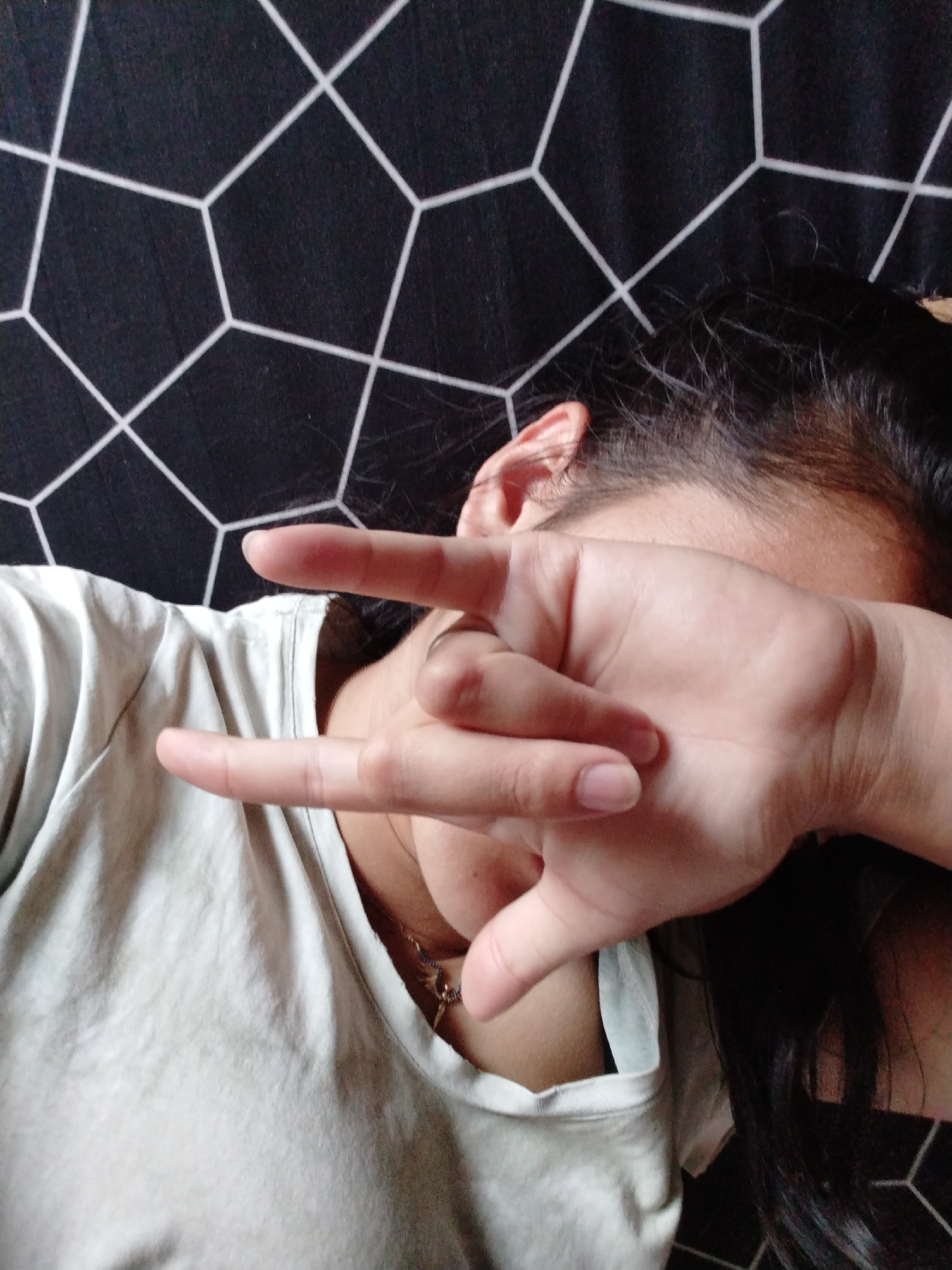 A person wearing a light-colored t-shirt lies on a surface with a black and white geometric pattern. Their right hand is raised, obscuring part of their face, with the index and pinky fingers extended. Part of an ear and dark hair are visible. A necklace hangs around their neck. The city and specific location are unknown.