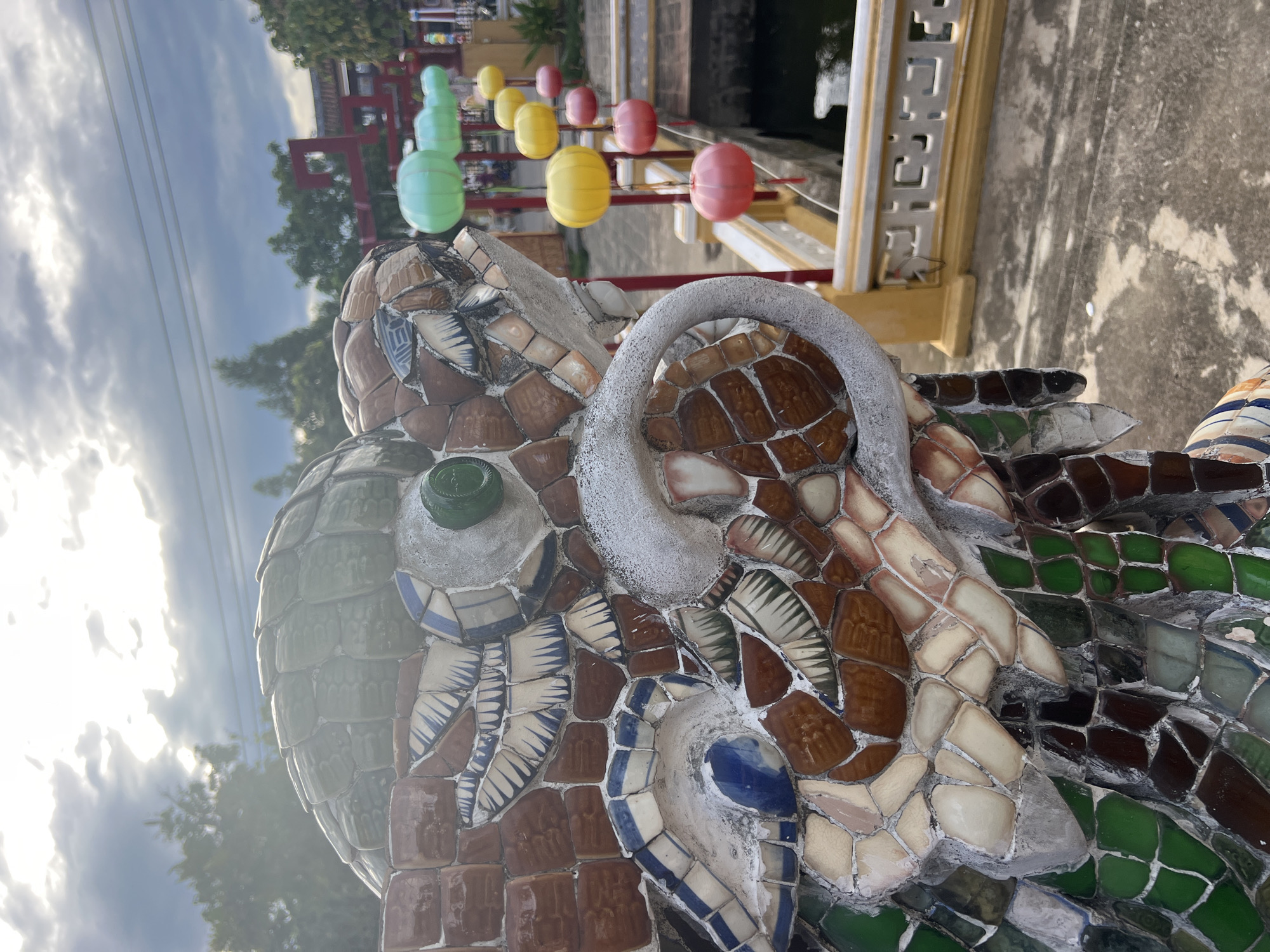 A close-up, eye-level shot captures a mosaic dragon statue in the foreground, its head and neck rendered in colorful ceramic shards of brown, white, blue, green, and reddish-orange. The dragon's snout is prominently featured, with a white, curved horn-like protrusion. Behind the dragon, a line of colorful, cylindrical lanterns in green, yellow, and red hangs suspended from poles, leading towards a traditional Vietnamese red gate structure in the distance. The lanterns are spaced evenly along a railing that borders a body of water, visible in the mid-ground. The sky above is overcast with patches of bright sunlight filtering through, casting a diffused light on the scene. The ground appears to be paved with concrete. The overall setting suggests a location within Hoi An, Vietnam, likely a temple or a historical site known for its decorative artistry and cultural ambiance.
