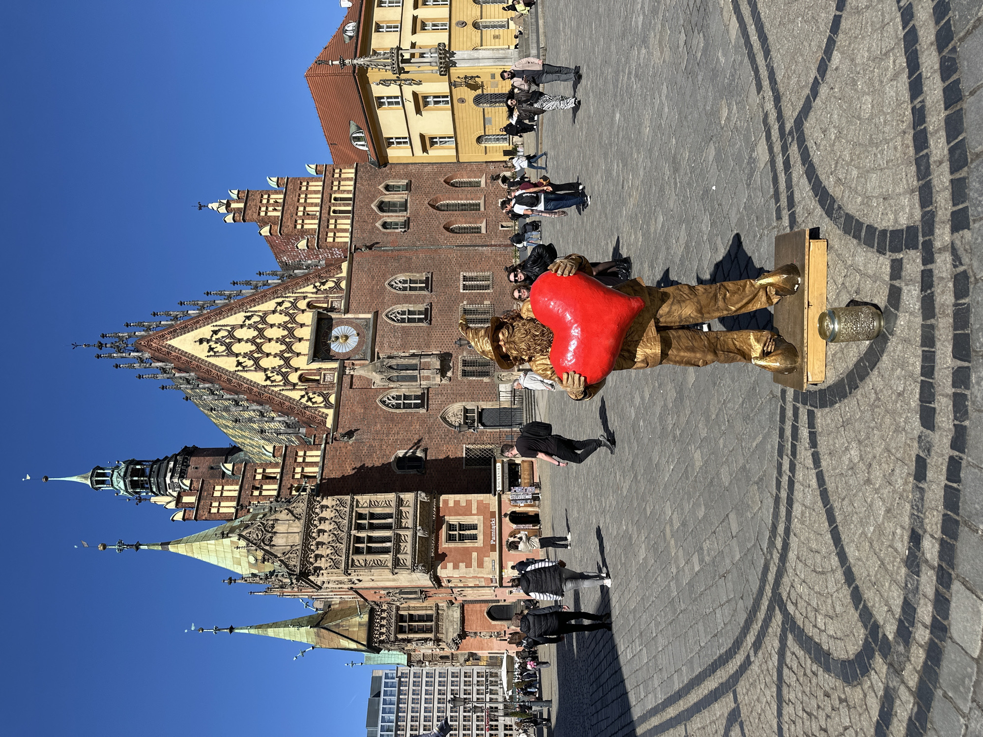 The image shows the Market Square (Rynek) in Wrocław, Poland. The Old Town Hall (Stary Ratusz) is prominent in the background. A street performer, painted gold and holding a large red heart, stands on a platform for tips. Several pedestrians are visible in the square. The sky is clear blue. The pavement is cobblestone.