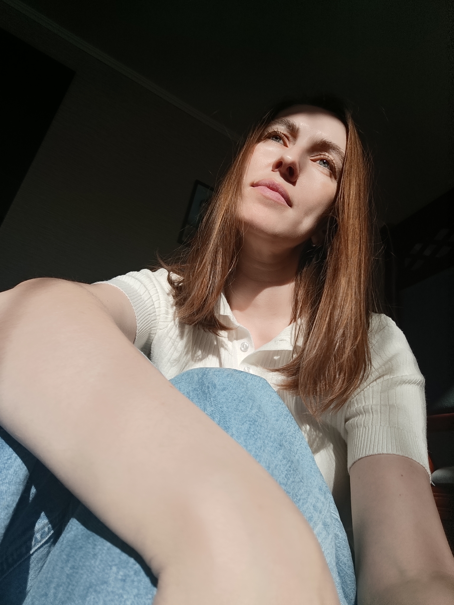 A woman with shoulder-length brown hair is sitting indoors, angled slightly upward and to the right. She's wearing a white, button-down shirt and blue jeans. Her left arm is in the foreground, resting on her leg. Her face is illuminated by sunlight, and she appears to be looking up and to the right. The background is dark, with a hint of wall details. The location is not determinable as Фоки, Russia, from the context given.