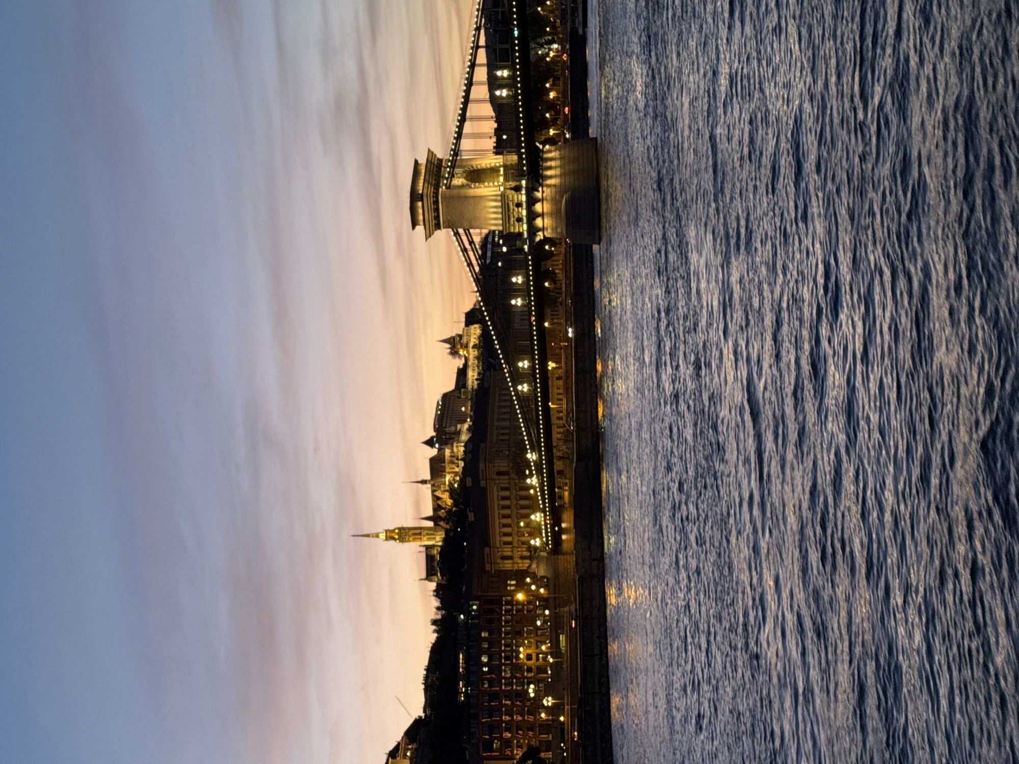The image shows the Chain Bridge in Budapest, Hungary, illuminated at twilight. The Danube River flows to the right. The Buda side skyline is visible, featuring various buildings and a church spire. The sky is a mix of blue and orange hues.