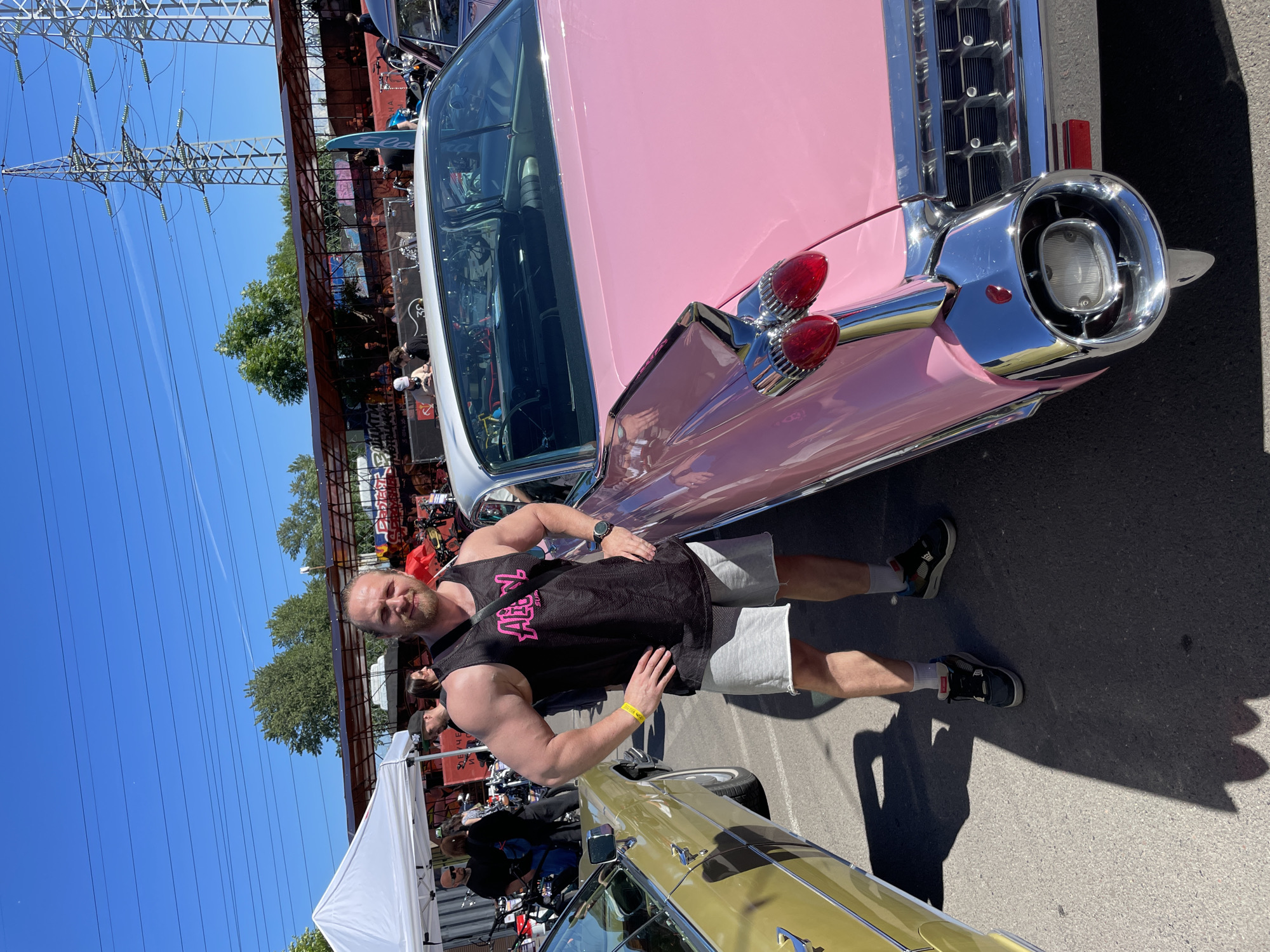 An adult male with light skin and brown hair is posing between two vintage cars on a paved lot under a clear blue sky. The man is wearing a black sleeveless tank top with pink lettering, grey athletic shorts, and black and grey sneakers. He has a muscular build and is standing with his hands on his hips, looking at the camera.
To his left is a yellow vintage car, and to his right is a pink vintage car with a white roof. The pink car features prominent chrome accents and distinctive tail fins with red taillights. In the background, there are tents, a structure with a red awning, and power lines and towers. Several other people and vehicles are visible in the background, suggesting an outdoor event.
The scene is likely at an outdoor car show or festival. No specific landmarks or architectural elements definitively place this image in Saint Petersburg, Russia, although the presence of overhead power lines and the general aesthetic are common in various urban and suburban settings.