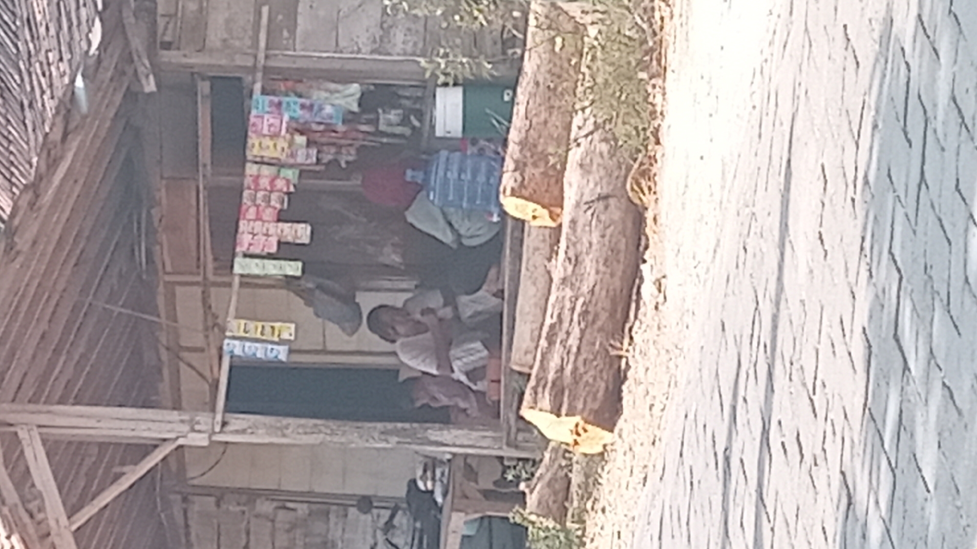In Kedungwaru, Indonesia, this image captures a scene in front of a rustic, open-air structure. Two individuals are seated on the ground under the eaves. The person on the left, wearing a light-colored shirt and striped pants, is holding a small, white object near their mouth. To their right, another person in a green shirt and red headscarf sits with their back to the camera, facing a large blue water cooler. In front of the structure, several large logs are laid out on the ground. A wooden table is visible to the far left. A clothesline strung with various small packaged goods hangs across the entrance. The ground in front of the scene is paved with interlocking bricks. The overall impression is one of a humble, possibly informal, roadside dwelling or shop.