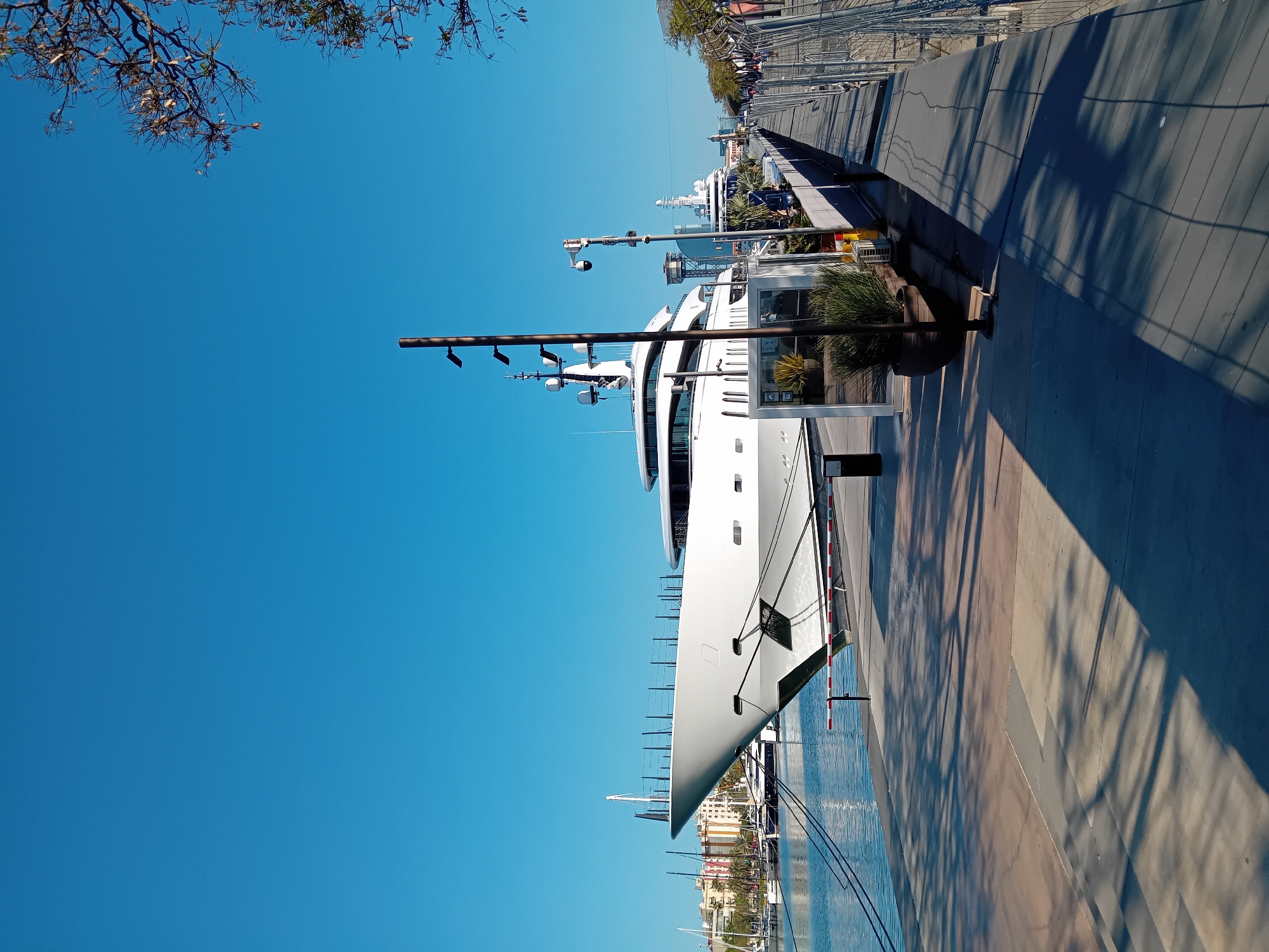 The image displays a large, modern white superyacht docked at a marina under a clear, bright blue sky, consistent with a sunny day in Barcelona, Spain. The yacht, featuring sleek lines and multiple decks adorned with antennas and radar equipment, occupies a significant portion of the right side of the frame.
A wide, paved promenade runs alongside the docked yacht, characterized by alternating sections of light and dark material. Several tall metal poles, some fitted with lights or cameras, are positioned along the promenade, casting long, distinct shadows across the ground, indicating either morning or late afternoon sunlight. In the water, to the left of the yacht's bow, reflections shimmer, and distant smaller sailboats are visible.
Further in the background, across the water, a cityscape with various buildings can be discerned. The upper left corner features the silhouette of bare tree branches against the vivid blue sky. Potted greenery is placed intermittently along the promenade near the yacht. The scene conveys a sense of tranquility and luxury, with no people explicitly visible. There is no legible text present in the image.