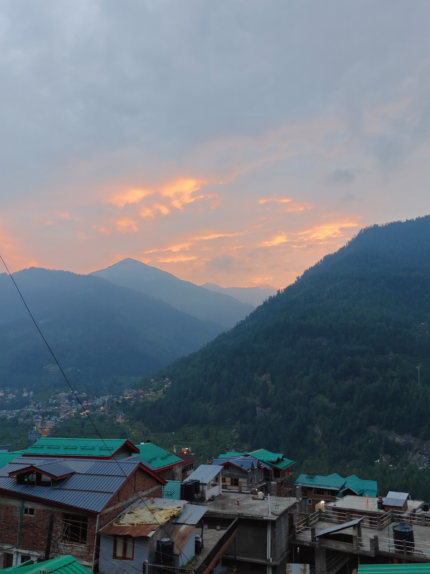 The image depicts a mountainous landscape at dusk, with a village nestled in the valley. The sky is a dramatic canvas of clouds, tinged with vibrant orange and yellow hues from the setting sun, contrasting with the otherwise overcast grey sky. Majestic, tree-covered mountains dominate the background, their slopes fading into a misty blue as they recede into the distance.
In the foreground, a collection of buildings with brightly colored, sloped roofs, predominantly green and some red, are visible. These structures appear to be part of a village or town, with some showing signs of wear and tear, like rusted metal sheeting. The architecture suggests a rustic, possibly residential or guesthouse setting. The overall scene evokes a sense of tranquility and the natural beauty of a mountainous region at the end of the day. The time of day is clearly evening or dusk, given the sky's coloration and lighting. The weather appears to be cloudy but not raining. The location context of Vashisht, India, aligns with the visual cues of mountainous terrain and distinct architectural styles often found in Himalayan regions. No text is visible in the image.
