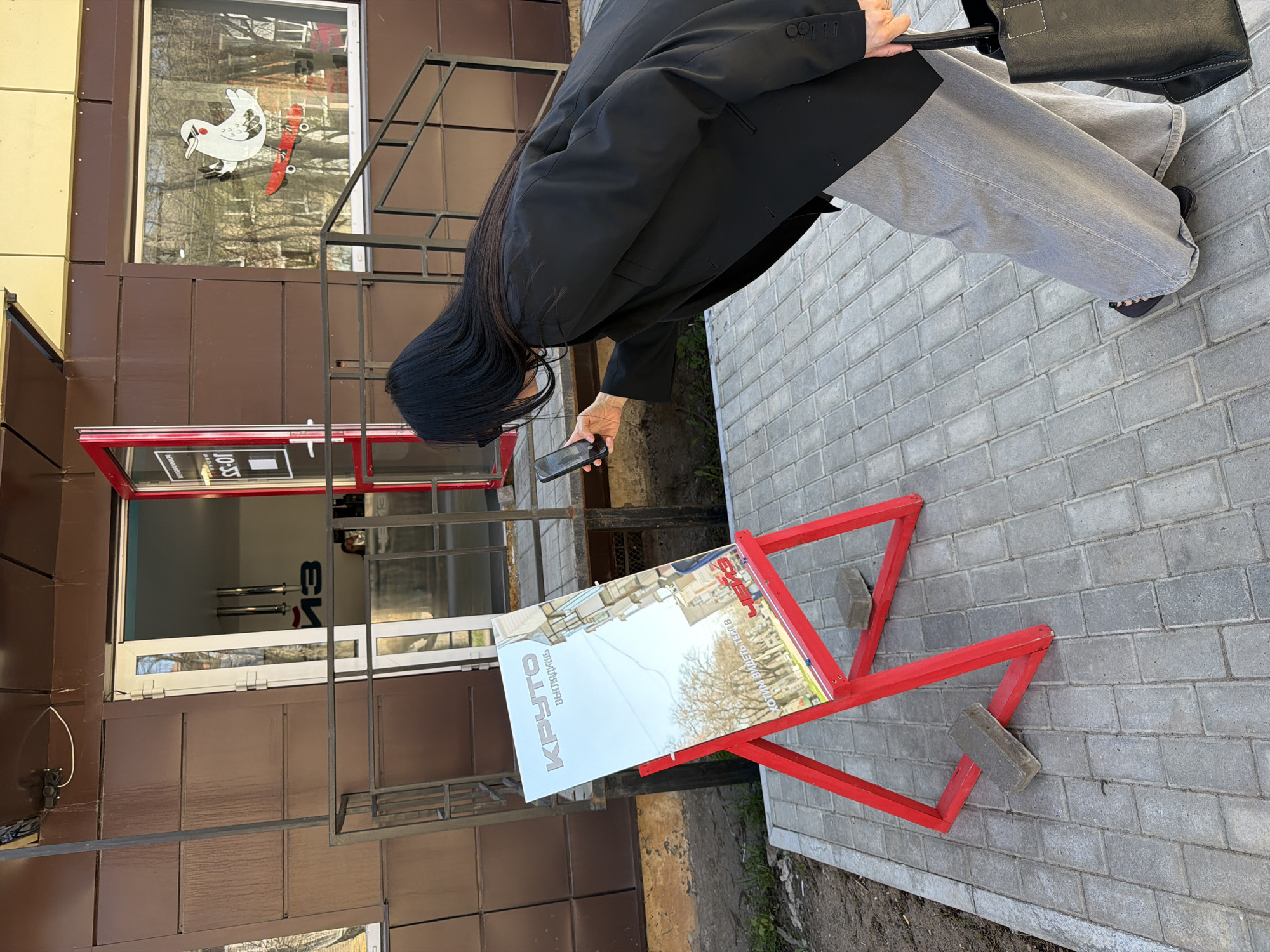 A person in a black blazer and gray pants is photographing a red, A-frame sign. The sign advertises "KRUTO" in Cyrillic script, located in the city of Chaikovsky, Russia. The sign is secured with gray bricks, placed on a gray brick sidewalk in front of a brown-paneled building with black metal trim. A window display shows a white bird on a skateboard. The person is holding a black smartphone. The person is also holding a black purse.