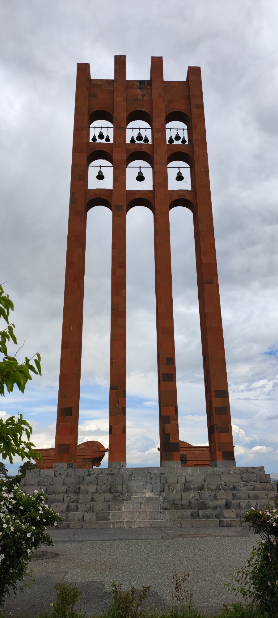 The image depicts a tall, three-part structure in the city of Araks, Armenia. It is constructed of reddish-brown stone with a pattern of darker blocks distributed throughout. The structure is composed of three parallel columns, connected by arched openings. Within the upper arches, black bells are suspended. The structure rests upon a wide, tiered stone base with two cannon replicas on top, near the base of the columns. The background consists of a cloudy sky with patches of blue visible through the structure's openings. In the foreground, there is vegetation, including flowers, partially obscuring the view of the base.