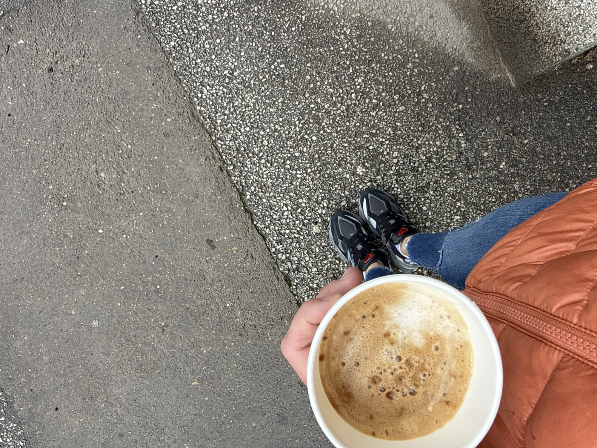 A first-person perspective photograph shows a person holding a cup of coffee while standing on a sidewalk. The person wears blue jeans and an orange puffer jacket, and black and gray running shoes with red accents. The ground is paved with two different types of concrete: one is smooth and gray, the other is rougher, with small stones embedded in the surface. The coffee in the white paper cup is light brown with a frothy top. The location cannot be definitively determined as being in Pleven, Bulgaria, based on the visual information provided.
