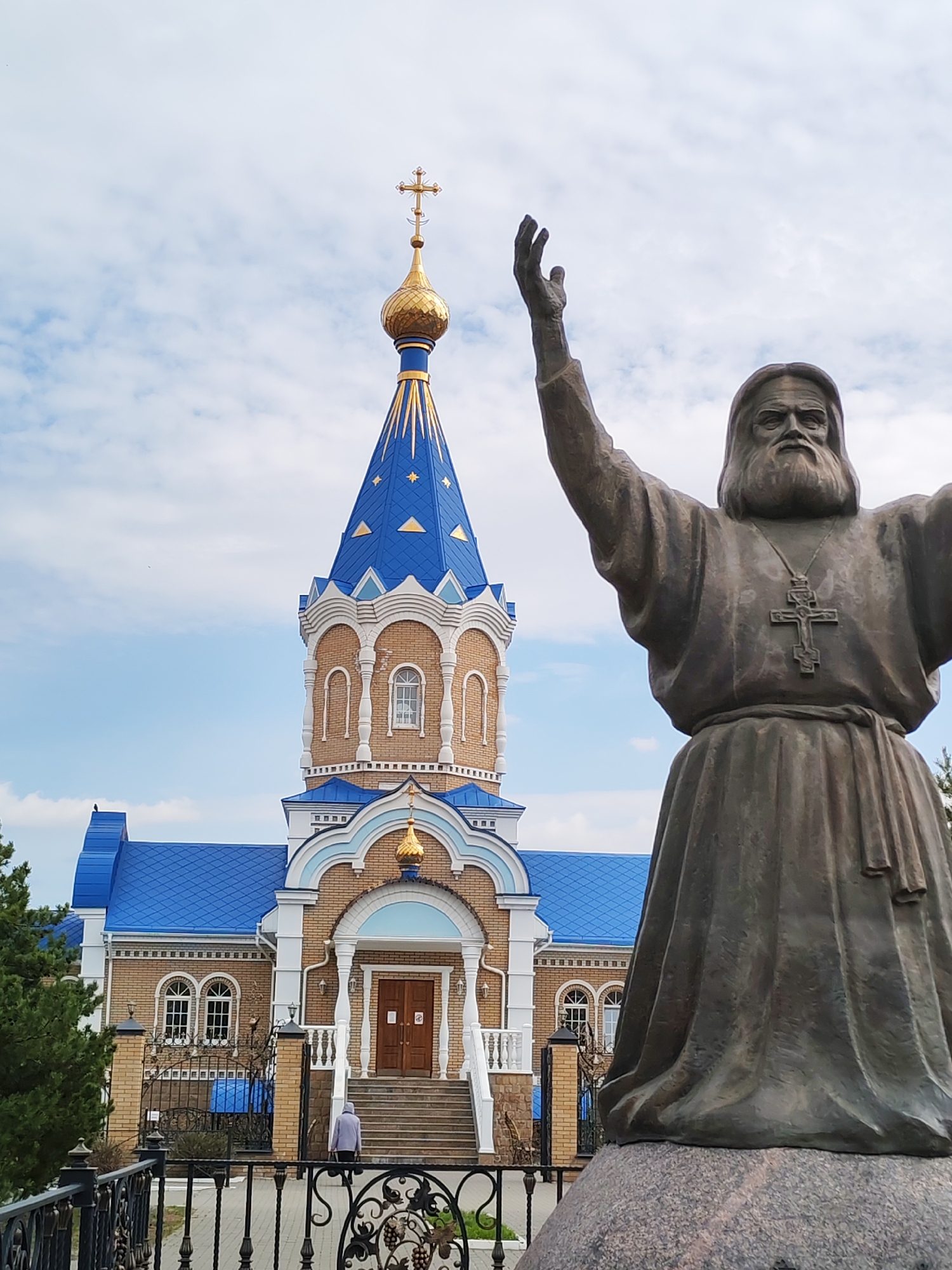 A bronze statue of a bearded man with a cross around his neck stands in the foreground, his arms raised in a gesture of blessing or supplication. In the background stands the Church of the Holy Grand Duchess Elizabeth in Izhevsk, Russia. The church features a blue roof, a tower topped with a golden dome, and white trim. The church is constructed of light-colored bricks. A black metal fence separates the viewer from the church grounds. A woman walks up the stairs towards the church's entrance. The sky is cloudy.