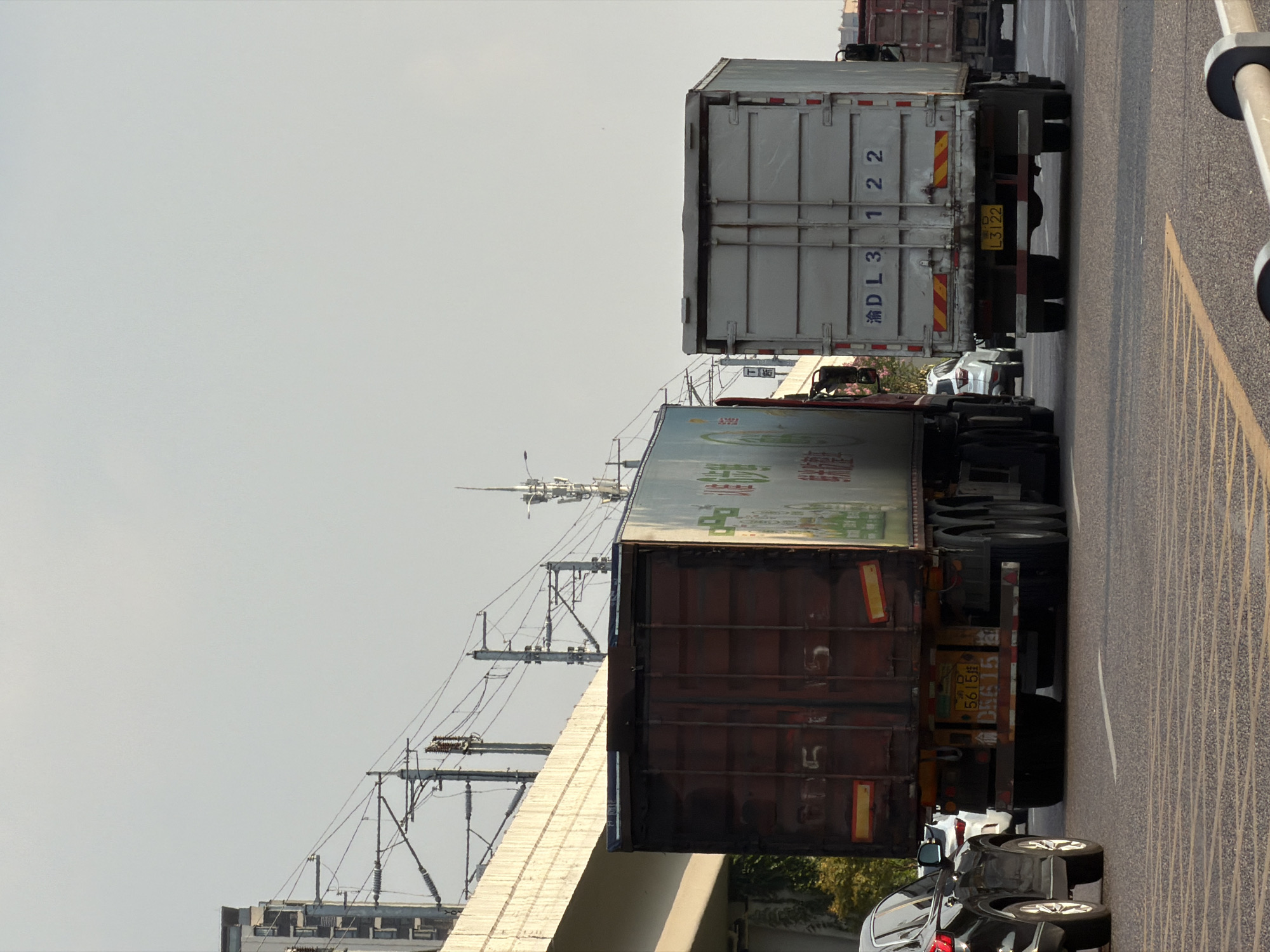 The image depicts a street scene in Jiangjin, China. Two large trucks are the prominent subjects, traveling on a multi-lane road. The truck on the left has a dark red cargo container with visible markings. The truck on the right has a white cargo container with blue lettering that reads "渝DL3 122". A black SUV is visible to the left of the trucks. In the background, elevated structures, possibly an overpass or railway, with electrical wires and poles are present. The sky is overcast. The road surface is asphalt with distinct lane markings.
