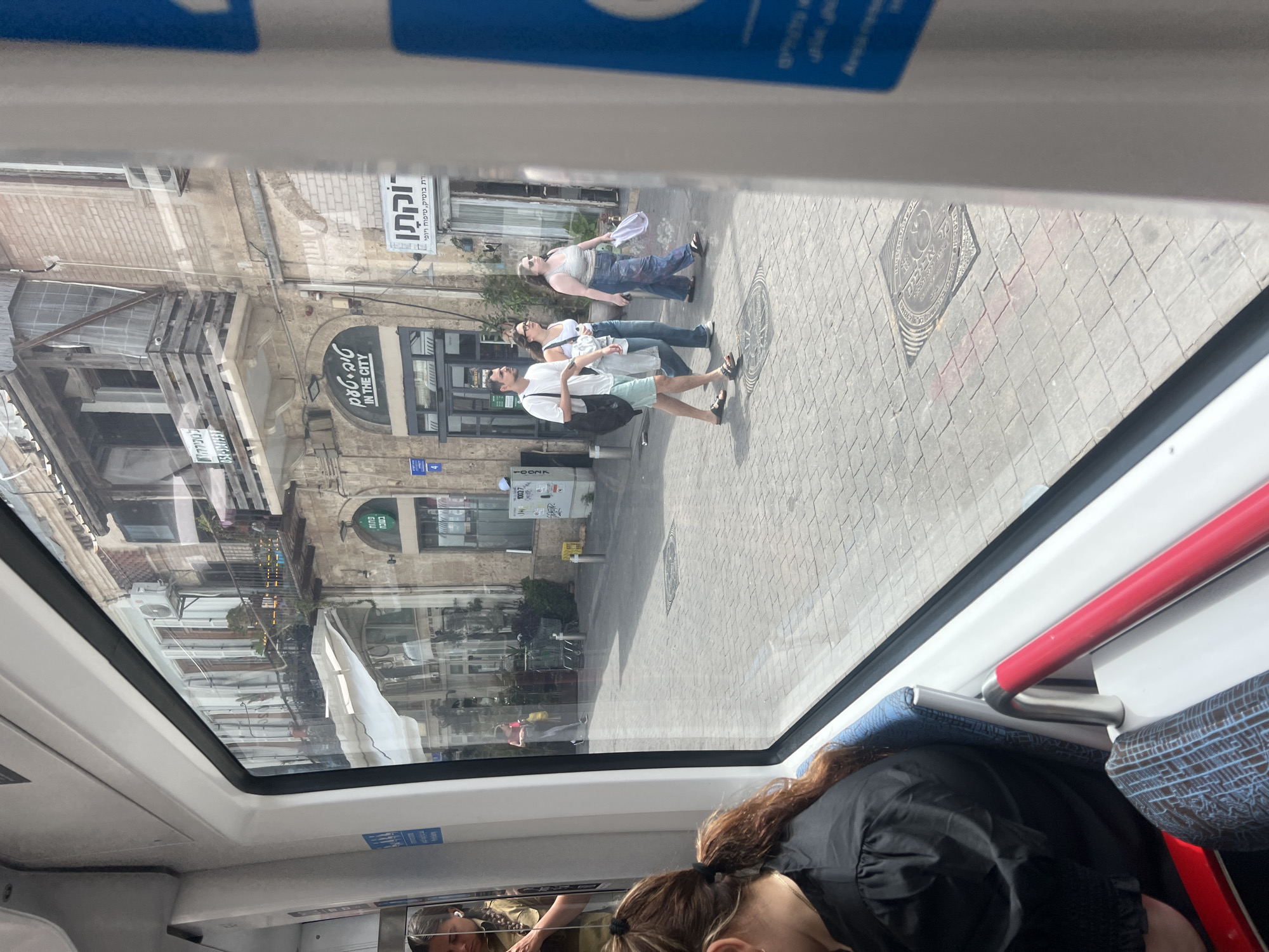 The image, taken from inside a vehicle, shows a street scene reflected in a window. Three people are walking on a brick-paved sidewalk: a man in a white shirt and light-colored shorts carrying a black bag, followed by two women wearing jeans and light-colored tops. Buildings line the street, including a store with a sign that says "IN THE CITY" and another with Hebrew writing. A manhole cover is visible on the sidewalk. The image includes reflections and views of the vehicle's interior, revealing a sleeping woman with brown hair and a person in the reflection of the window. The location is on Nahalat Binyamin Street in Tel Aviv, Israel.