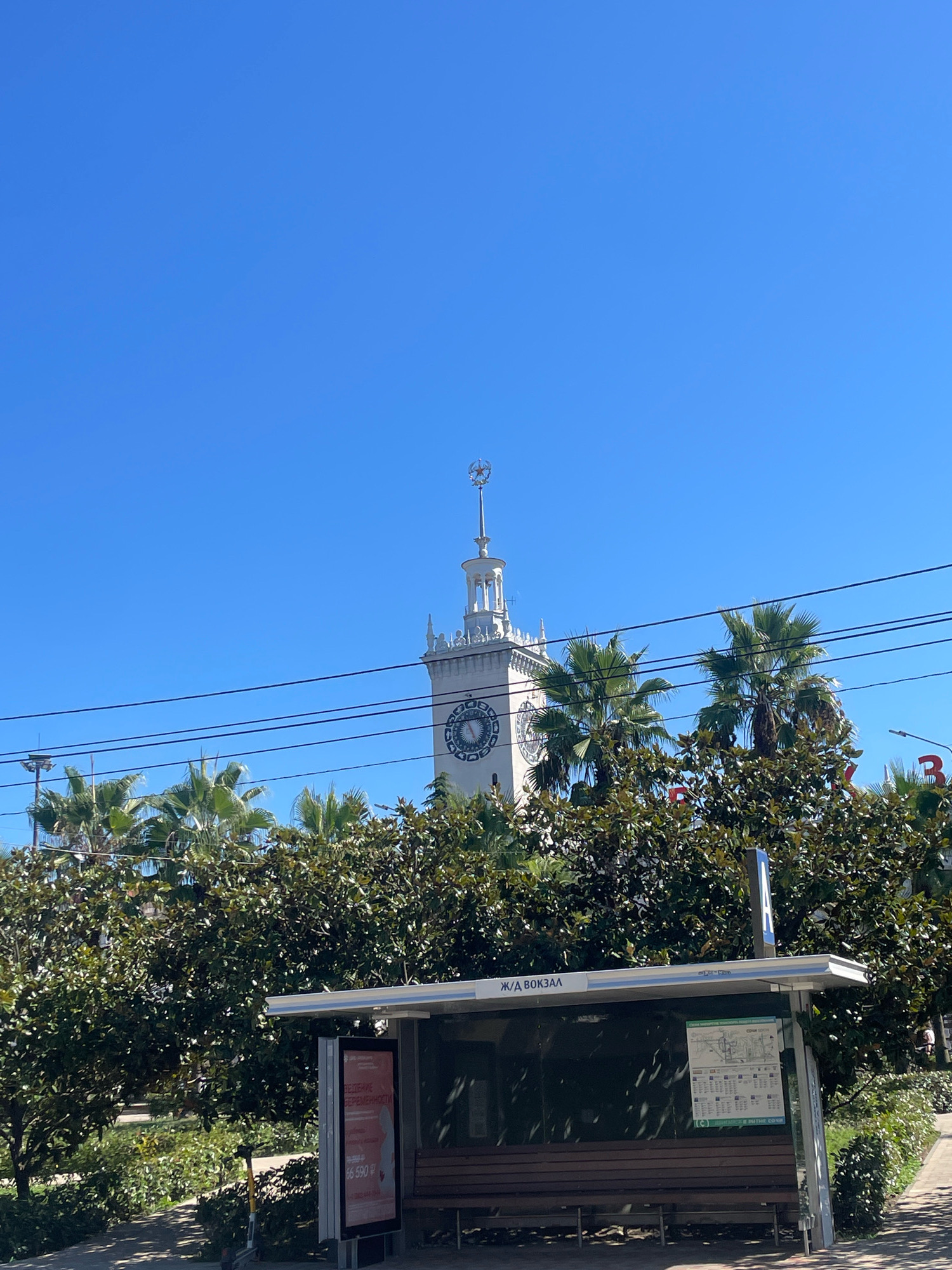 The image depicts a clear, sunny day in Sochi, Russia, with a bright blue sky dominating the upper portion. In the background, a distinctive white clock tower with a star ornament at its apex rises above lush green trees, including palm trees. The tower has a large clock face with Roman numerals. In the foreground, a bus stop shelter is visible. The shelter has a transparent roof and sides, with a wooden bench inside. A sign on the shelter reads "Ж/Д ВОКЗАЛ," indicating it is a train station bus stop. An advertisement is displayed on a panel within the shelter, featuring text and graphics, including a price of "66 590 ₽". Adjacent to the shelter, a light-colored electric scooter is parked on a paved pathway. The pathway is lined with green bushes and trees. Overhead power lines stretch across the image, partially obscuring the view of the clock tower and palm trees. To the right of the bus stop, a lamppost is visible, and beyond that, a red sign with the letter "Б" is partially visible.