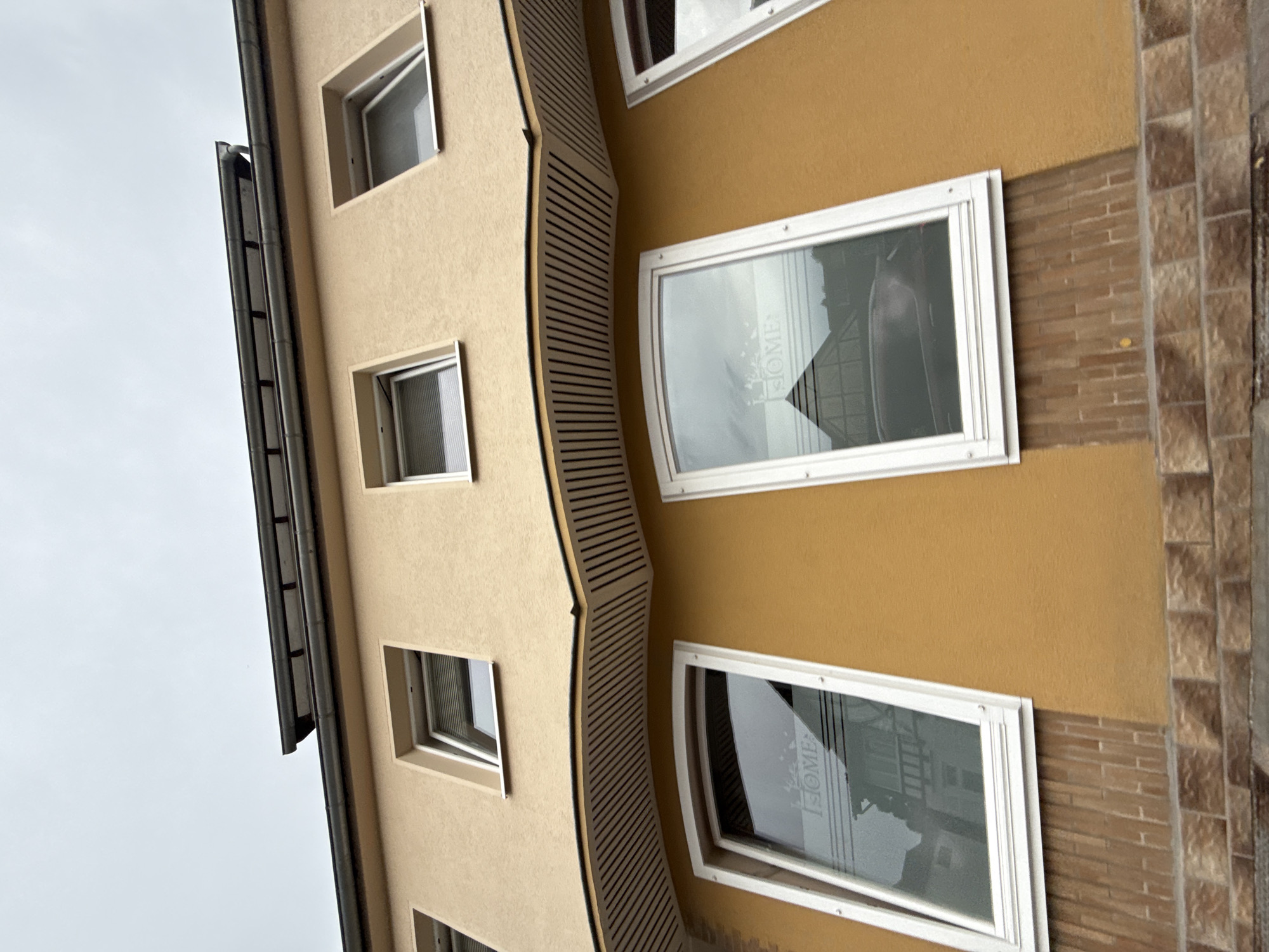 The image shows the exterior of a building in Bad Sachsa, Germany, on a cloudy day. The building has a light yellow facade with several windows on two levels. The upper level has smaller, square windows, some with blinds. The lower level features larger, rectangular windows with white frames. One of these lower windows has text that appears to read "HOME" reflected on its surface. Below the lower windows, there is a section of brickwork, and the building rests on a foundation of stone blocks. A dark, wavy soffit with parallel lines is visible between the two levels of windows. The roofline is visible at the top, with what appears to be solar panels installed. The overall impression is of a residential or commercial building.
