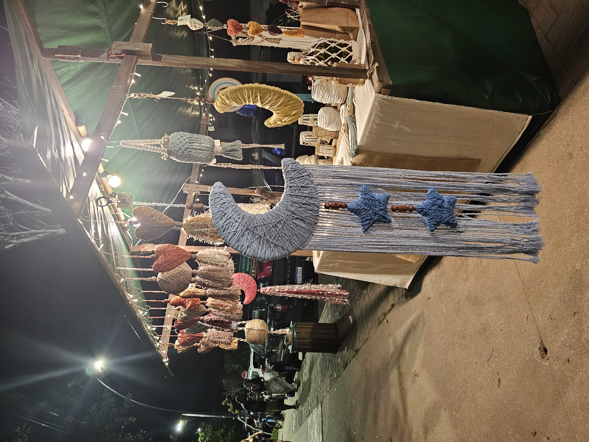 The image depicts an outdoor craft market stall at night in Armação dos Búzios, Brazil. The stall is constructed from a rustic wooden frame, covered by a dark green tarp-like roof, and illuminated by numerous string lights, creating a warm ambiance.
The primary subjects are various handmade decorative items, predominantly crafted from yarn and natural fibers. In the foreground, a large, light blue yarn-wrapped crescent moon hangs, accompanied by two smaller blue yarn stars, suspended from strands of matching blue yarn. To the left, an extensive collection of colorful tassels and pom-poms, in shades of red, orange, beige, and blue, are displayed hanging from horizontal wooden rods. Another notable hanging item is a textured yellow crescent shape, possibly also made of yarn. Other woven and wrapped decorative objects are visible hanging or arranged on tables within the stall.
The activity taking place is an evening market or craft fair, where artisans are showcasing and selling their handmade goods. In the background, beyond the brightly lit stall, the scene is dark, punctuated by distant streetlights. Several cars are parked, including one with the license plate "KKW2A03." Indistinct figures of people can be seen, suggesting market-goers or vendors.
Notable details include the time of day, which is clearly night due to the dark sky and reliance on artificial lighting. A street sign in the lower-left background clearly reads "PARE" (Portuguese for "STOP"). Another partially obscured sign in the mid-ground shows "Alternativ" and "GE". The overall atmosphere is cozy and inviting, highlighting local craftsmanship.