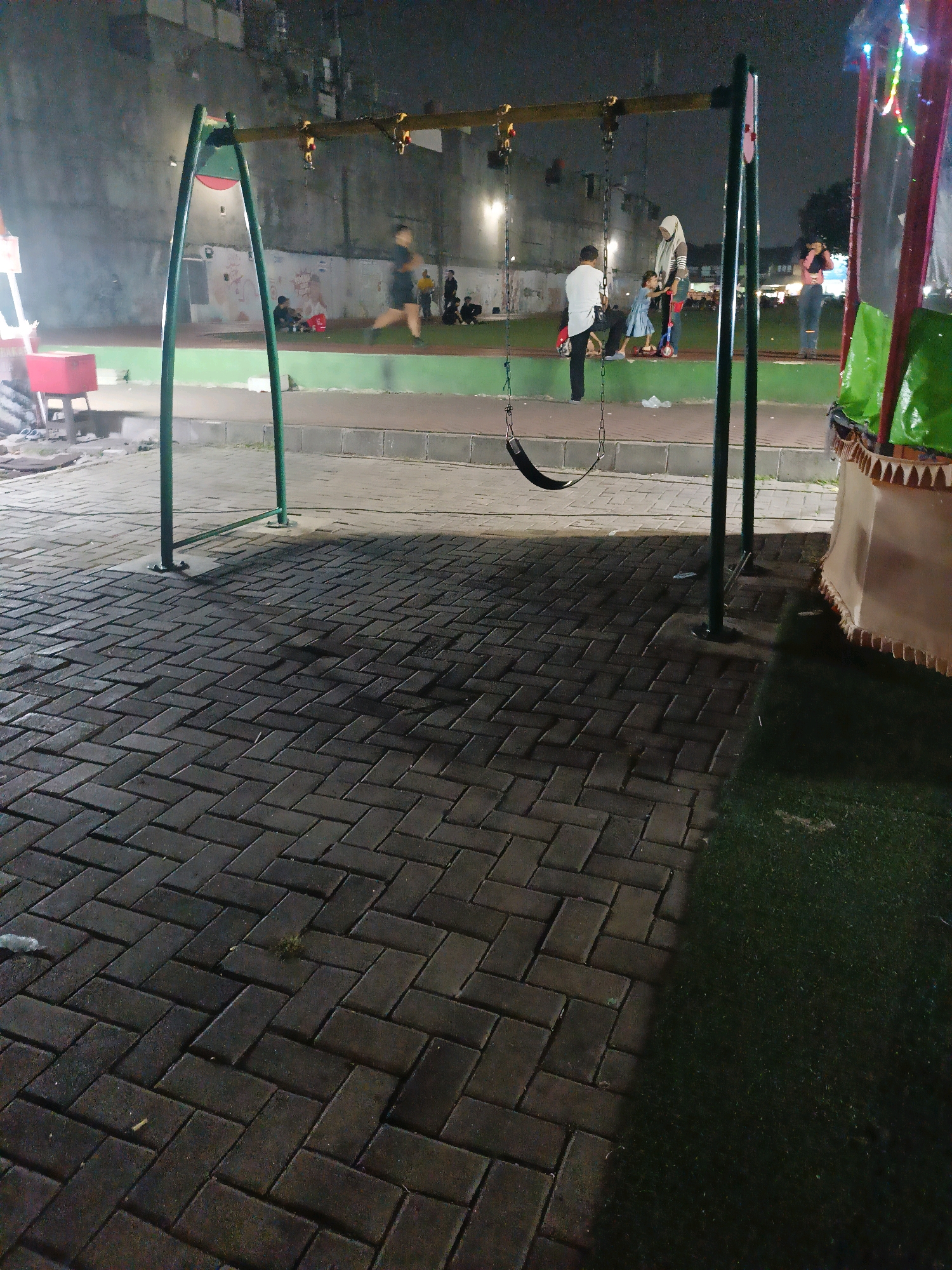 A nighttime scene depicts an outdoor playground area paved with dark interlocking brickwork. A swing set with a single black swing is the prominent feature in the foreground. In the background, several individuals are present. A person in a white shirt and black pants stands near a young child on a scooter. A woman wearing a hijab is also present, interacting with the child. Further in the background, other individuals are visible, some appearing to be standing or sitting. The environment includes a concrete wall, a grassy area, and what appears to be part of a structure with green and brown fabric at the far right. The lighting suggests artificial illumination from the playground and possibly nearby sources.