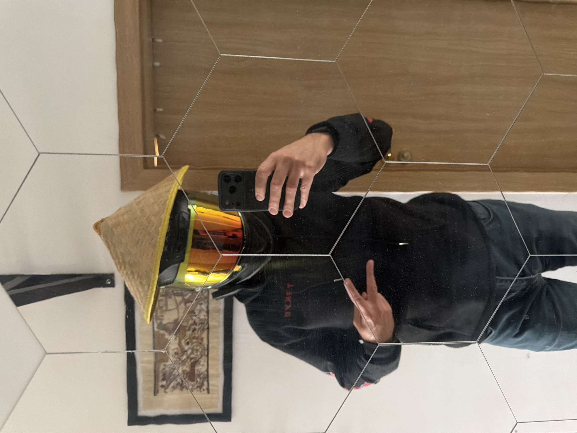 The image shows a person's reflection in a mirror comprised of hexagonal tiles. The person is wearing a black hoodie with red lettering, blue jeans, and a motorcycle helmet with yellow goggles. They are also wearing a conical Asian hat. The person is holding a smartphone and making a peace sign with their hand. The reflection includes a framed artwork and a wooden door frame. There is no indication in the image that it is located in Sofia, Bulgaria.