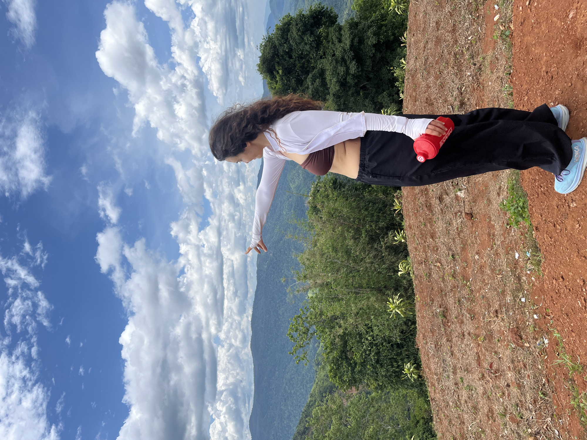 In Nao Sẻ, Vietnam, a woman stands on a reddish-brown dirt hill, pointing towards a vast expanse of lush green mountains and rolling hills under a bright blue sky dotted with fluffy white clouds. She has long, dark, curly hair and wears a brown bikini top, a white long-sleeved shrug, and black loose-fitting pants. She is holding a red water bottle in her right hand. The terrain in front of her appears to be recently cleared, with patches of dry grass and scattered debris. The background features dense vegetation, including bamboo and broad-leafed trees, receding into the distant, mist-covered mountain ranges. The overall scene suggests an outdoor activity like hiking or exploring in a natural, scenic setting.
