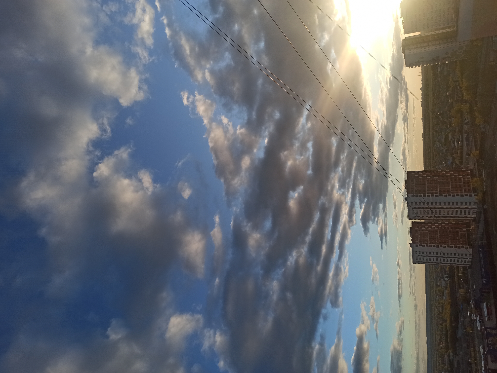 The image depicts a partly cloudy sky over an urban area. Power lines run diagonally across the frame. Buildings are visible on the right side of the image, including several high-rise apartment buildings. The image appears to be taken during sunrise or sunset, as the sun is low in the sky and the light is warm. Based on the location in the prompt, it is assumed that this scene is in Кондратово, Russia.