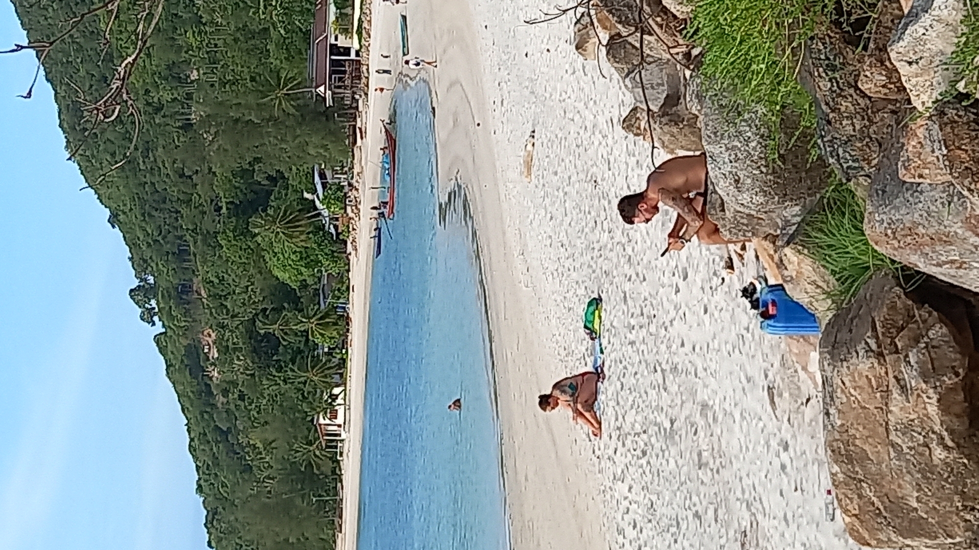 The image depicts a beach scene on the island of Koh Phangan, Thailand. In the foreground, a man is seated on large rocks, looking at his phone. To his left, a blue plastic container and a pair of shoes are visible. Further up the beach, a woman is sitting on the sand next to a green bag. In the water, a single person is swimming. The background features a sandy beach, the blue ocean, and a lush green hillside with scattered buildings and palm trees. Several boats are anchored in the shallow water. The sky is clear and blue.