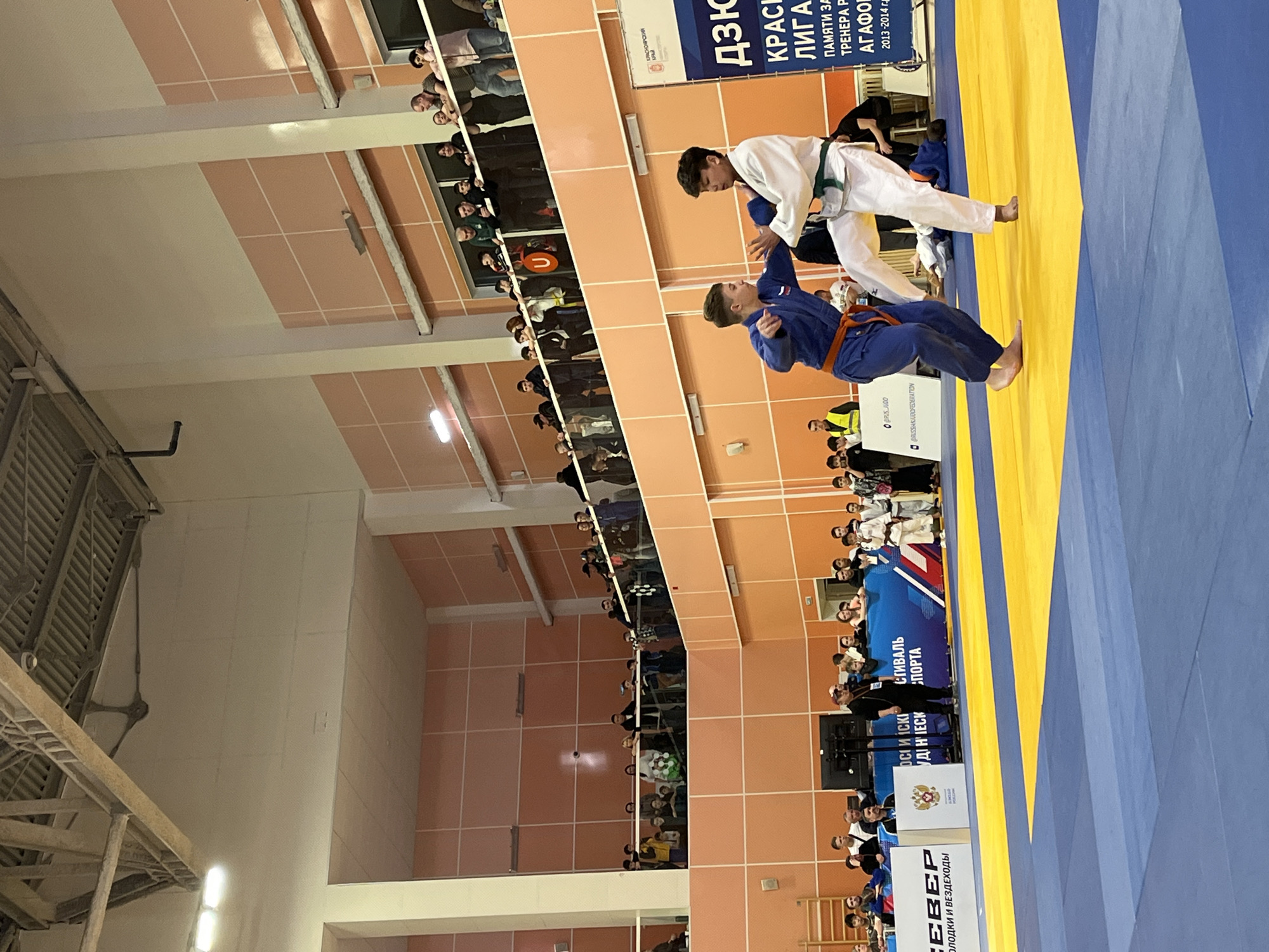 The image shows two judokas in action on a blue and yellow mat during a competition. One judoka is wearing a white gi with a green belt, while the other is wearing a blue gi with an orange belt. The judoka in white appears to be executing a throwing technique on the judoka in blue.
The competition is taking place indoors, with spectators in tiered seating along one side of the venue. Banners and signage indicate that the event is a Judo tournament in Krasnoyarsk, Russia. One banner mentions the "Красноярский Лига" (Krasnoyarsk League) and is dedicated to the memory of coach Agafonov (Агафонов).
Additional banners display logos for the Russian Judo Federation and other sponsors, including a company involved with youth sports or off-road vehicles ("молодки и вездеходы"). The venue's walls are tiled in peach and white. The lighting appears to be standard indoor sports facility lighting.