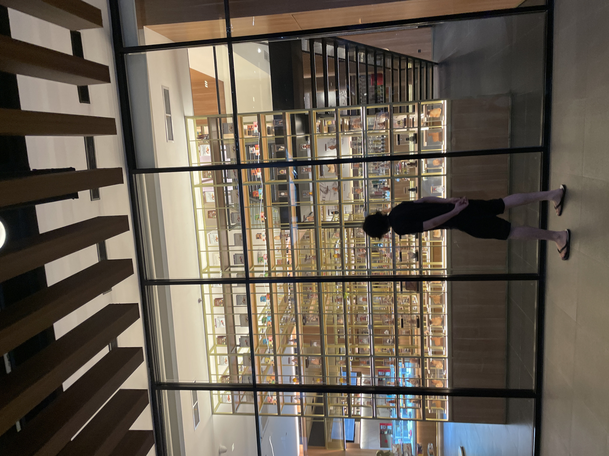A person is standing in front of a large glass display case in a well-lit, modern interior. The display case contains numerous shelves filled with a variety of items. The location appears to be in North Jakarta, Indonesia. The person is wearing dark clothing and sandals. The shelves are arranged in a grid pattern. Above the display case are dark, horizontal beams.