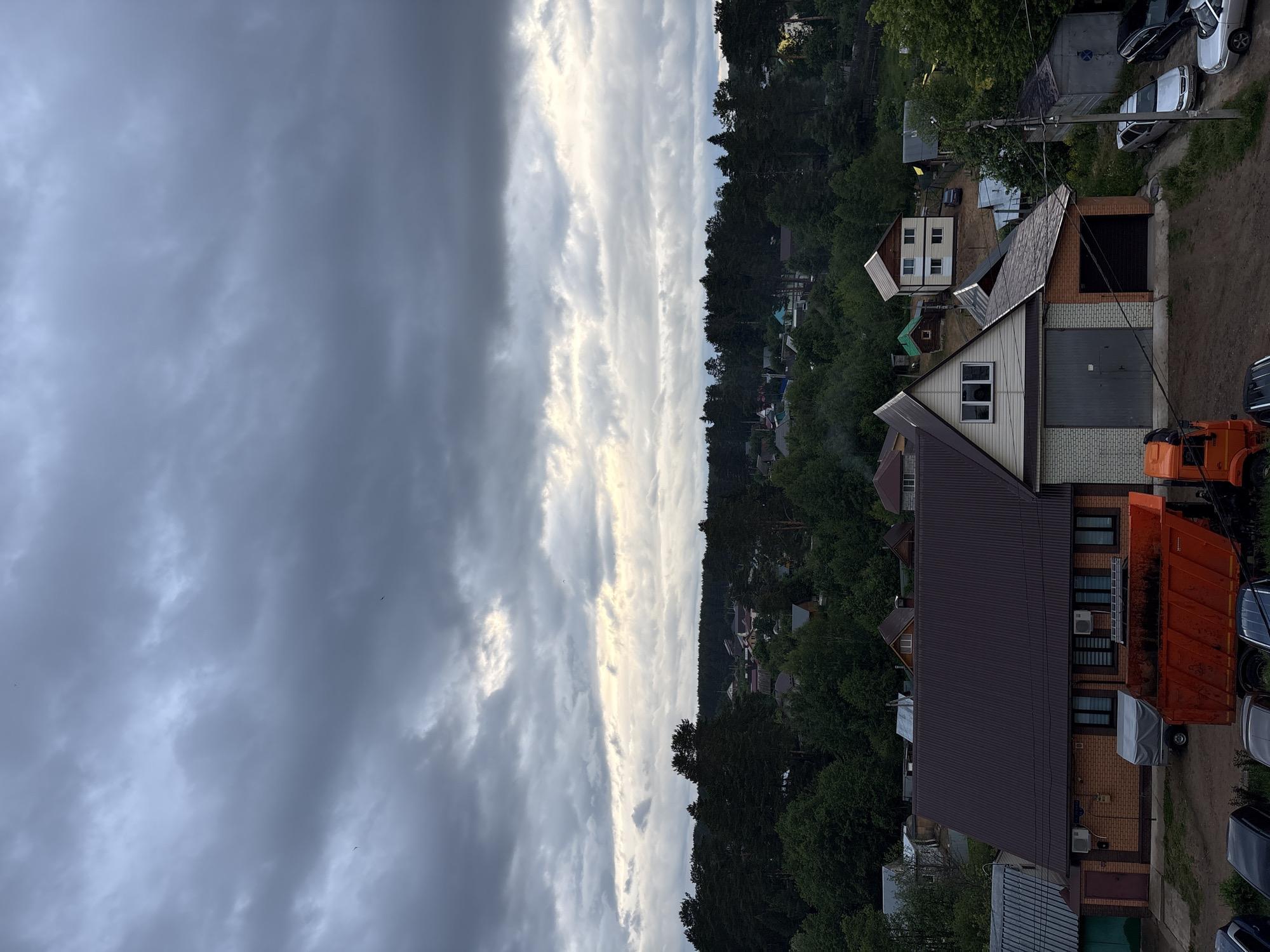 This is a high-angle view of a residential area within the city of Chaikovsky, Russia, characterized by numerous houses nestled amidst dense woodland under an overcast sky.
The upper two-thirds of the image are dominated by heavy, dark gray stratocumulus clouds, indicating overcast weather conditions. A narrow, lighter band of sky with faint hints of pale yellow light is visible above the distant horizon, particularly towards the right. Several small, dark avian silhouettes are present in the sky.
The midground features a vast, dark green expanse of forest, primarily comprising coniferous trees, forming a continuous horizontal band across the image. Scattered within and along the edges of this forest are numerous detached houses, varying in size and architectural style, indicating a low-density residential or dacha settlement.
The foreground presents closer views of several buildings and parked vehicles on unpaved ground.
On the left, a two-story building with light brown brick walls and a dark brown corrugated metal gable roof is prominent. It features multiple rectangular windows with dark frames and internal blinds, along with two exterior air conditioning units mounted on its facade. A dark red door is visible on its left side. A gray, tarp-covered trailer is parked in front of this building.
To the right, another multi-level building features a gabled roof and light-colored horizontal siding on its upper facade, including a central window unit. The lower level consists of a large gray roll-up garage door and an adjacent section of brickwork with a smaller dark roll-up garage door.
A large, orange dump truck with an empty, dark orange bed is conspicuously parked in the lower center of the image, facing left.
Several passenger vehicles are parked on the unpaved dirt areas: parts of dark-colored cars are visible on the far left, and on the right side, a silver sedan, a dark sedan, and another silver sedan are parked along a dirt track.
A small, rectangular utility shed with a dark, sloped roof is situated on the far right. A wooden utility pole with overhead wires is present on the right, positioned between the parked cars and the shed.
The ground throughout the foreground is unpaved, consisting of light brown, exposed soil. Abundant green foliage, including both deciduous and coniferous trees and shrubs, surrounds the buildings and fills the spaces between them. No people are visible in the scene.