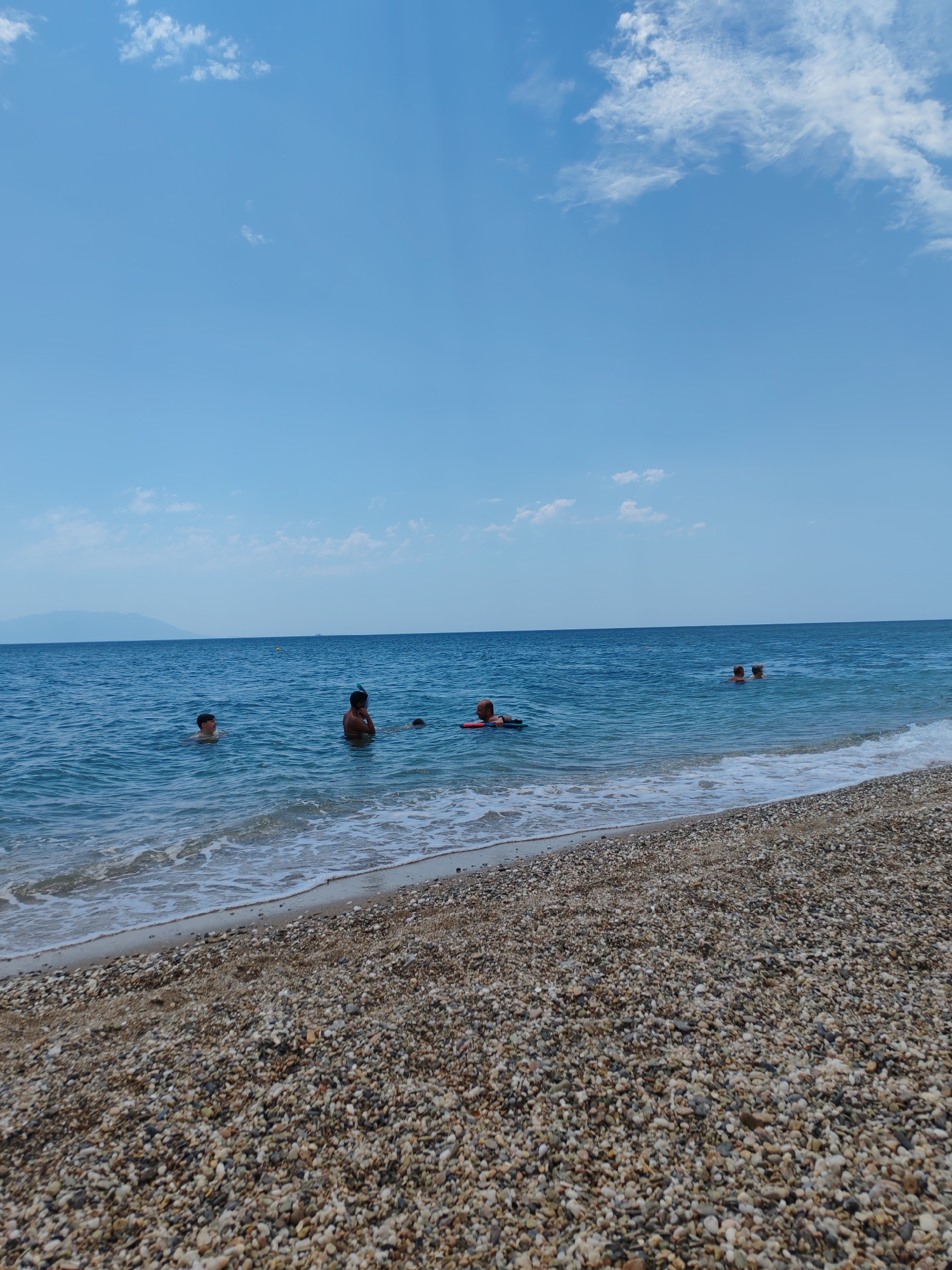 The image depicts a sunny day at a pebble beach in Makri, Greece. Several people are enjoying the water. In the foreground, a man is lying on a red flotation device. Further out in the sea, a young person with a snorkel mask is visible, as are two other individuals swimming. In the distance, a hazy landmass can be seen, likely an island or coastline. The sky is a clear blue with scattered white clouds, indicating pleasant weather. The sea is calm with gentle waves lapping the shore. The beach itself is covered in a variety of small, smooth pebbles. The overall scene suggests a relaxed and enjoyable day at the beach. No visible text is present in the image.