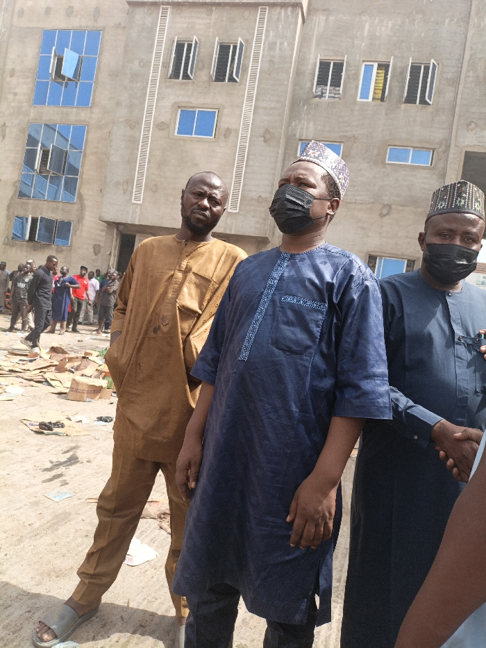 The image displays an outdoor scene in Fagge C, Nigeria, under daylight conditions. Three men stand prominently in the foreground against the backdrop of a multi-story building. The building exhibits an unpainted concrete facade featuring various window types: some are fitted with blue reflective glass, others with white frames and clear panes, and several remain open. Vertical decorative grooves are integrated into the building's exterior.
The ground area in the foreground and mid-ground is rough and unpaved, covered with scattered cardboard sheets and other debris.
From left to right, the three main subjects are:
1. A man with a bald head and short beard, wearing a light brown traditional tunic and trousers, and light-colored open-toe sandals. His hands are in his pockets, and he looks towards the camera with a neutral expression.
2. A man in a dark blue traditional outfit with light blue embroidery, a black face mask, and a patterned cap (kufi style). He is looking slightly upwards and to the right, away from the camera.
3. A man, partially visible on the far right, also in a dark blue traditional garment, a black face mask, and a patterned cap. His right hand is engaged in a handshake with an individual largely outside the frame.
In the mid-ground, behind the main subjects, a crowd of approximately 10-15 individuals, predominantly men in various casual and traditional attire, are gathered or walking near the base of the multi-story building. No specific interactions among this background group are clearly discernible.