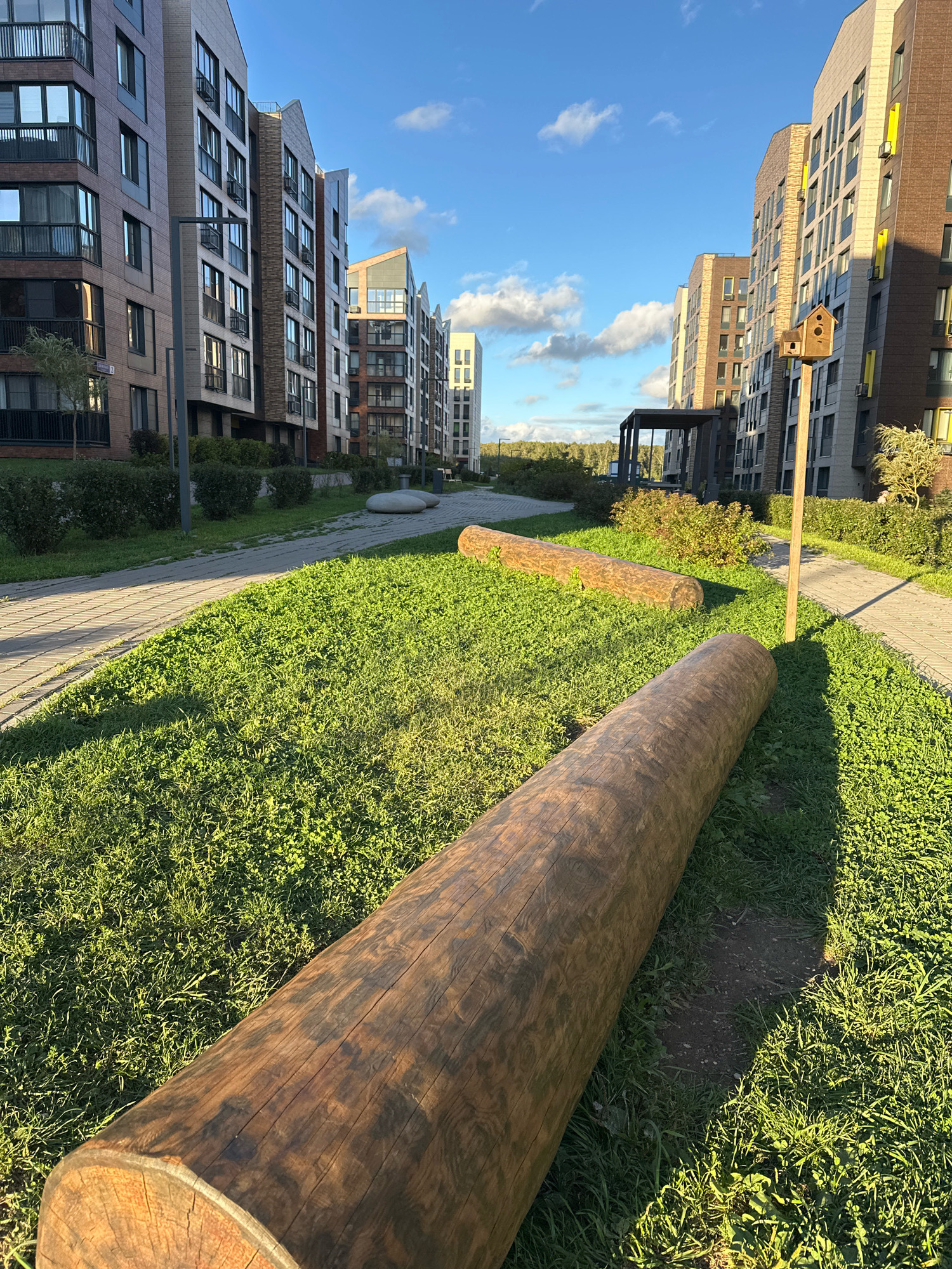 The image depicts an outdoor courtyard area in the city of Vidnoye, Russia, characterized by modern residential buildings and a landscaped environment. In the foreground, two large, fallen logs lie on a grassy area, serving as natural seating or decorative elements. A paved pathway curves through the scene, bordered by well-maintained green grass and low-lying bushes. In the mid-ground, the pathway continues, leading towards a covered, open-air structure. Further back, several multi-story residential buildings with contemporary architectural designs are visible, featuring a mix of brick and facade materials, with balconies and numerous windows. A wooden birdhouse is mounted on a pole next to one of the buildings. The sky is blue with scattered white clouds, indicating clear weather. The overall scene conveys a sense of modern urban living integrated with natural elements. There are no people or animals visible in the image.