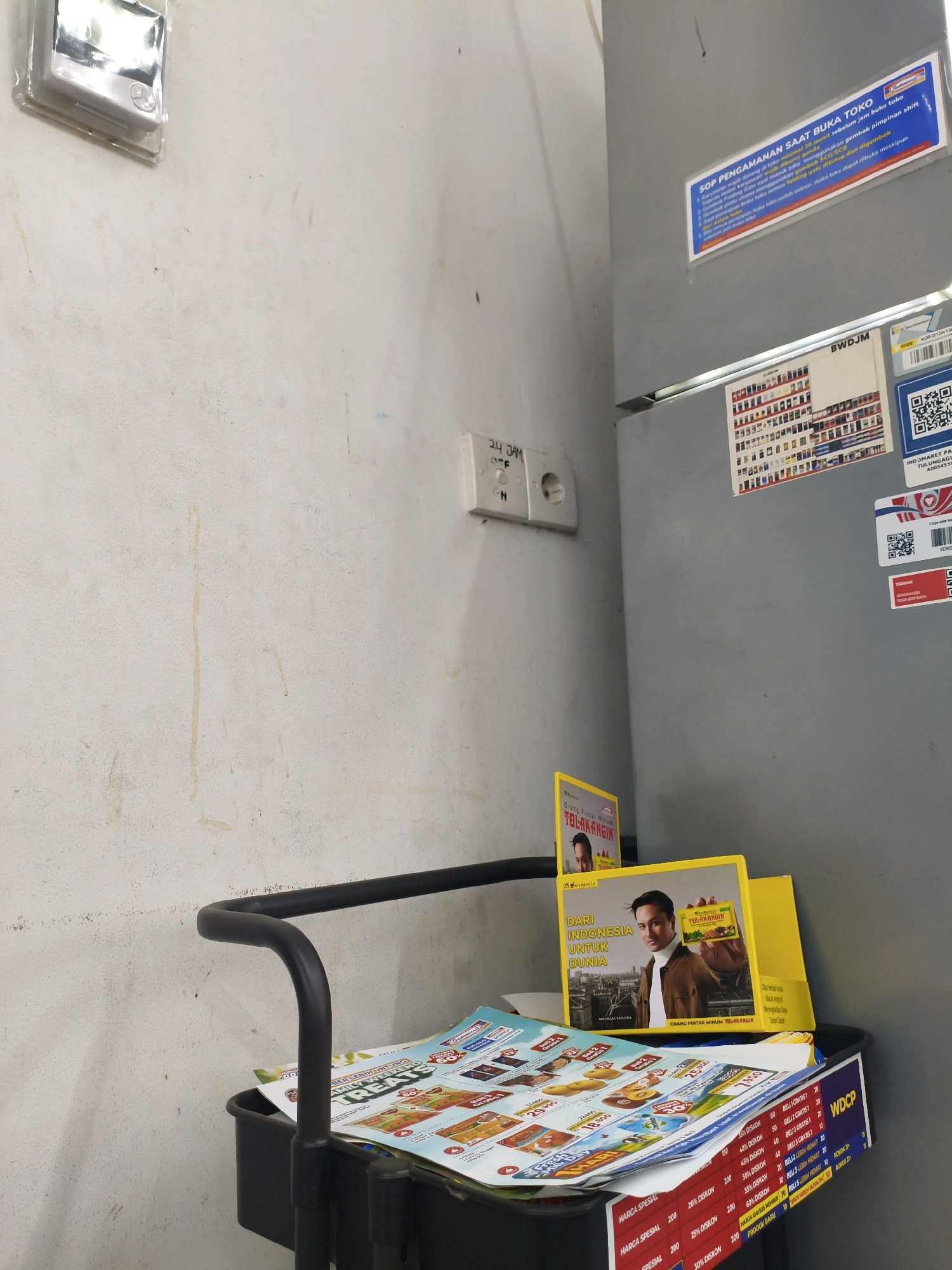 The image depicts an indoor scene, likely a retail environment, given the presence of promotional flyers and signage. The walls are textured and off-white, with some visible smudges and stains. On the left wall, a white electrical outlet labeled "24 JAM" (24 Hours) is visible.
Dominating the right side of the image is a grey, metallic surface, possibly a refrigerator or a display unit. Several stickers are affixed to this surface. One prominent blue and white sticker reads "SOP PENGAMANAN SAAT BUKA TOKO" (Security SOP for Store Opening), detailing procedures in Indonesian. Below this, a smaller white sticker displays "BWDJM" and a grid of product images. Further down, a QR code and text in Indonesian are visible.
In the foreground, a black cart holds a stack of promotional flyers. The flyers display colorful advertisements with text in Indonesian, featuring discounts and product images. One flyer prominently shows a man holding a yellow box, with text that includes "TOLAK ANGIN" (a traditional Indonesian herbal medicine) and "DARI INDONESIA UNTUK DUNIA" (From Indonesia for the World).
The lighting in the scene is neutral, casting soft shadows. The overall impression is one of a functional, possibly small, retail space in Indonesia.