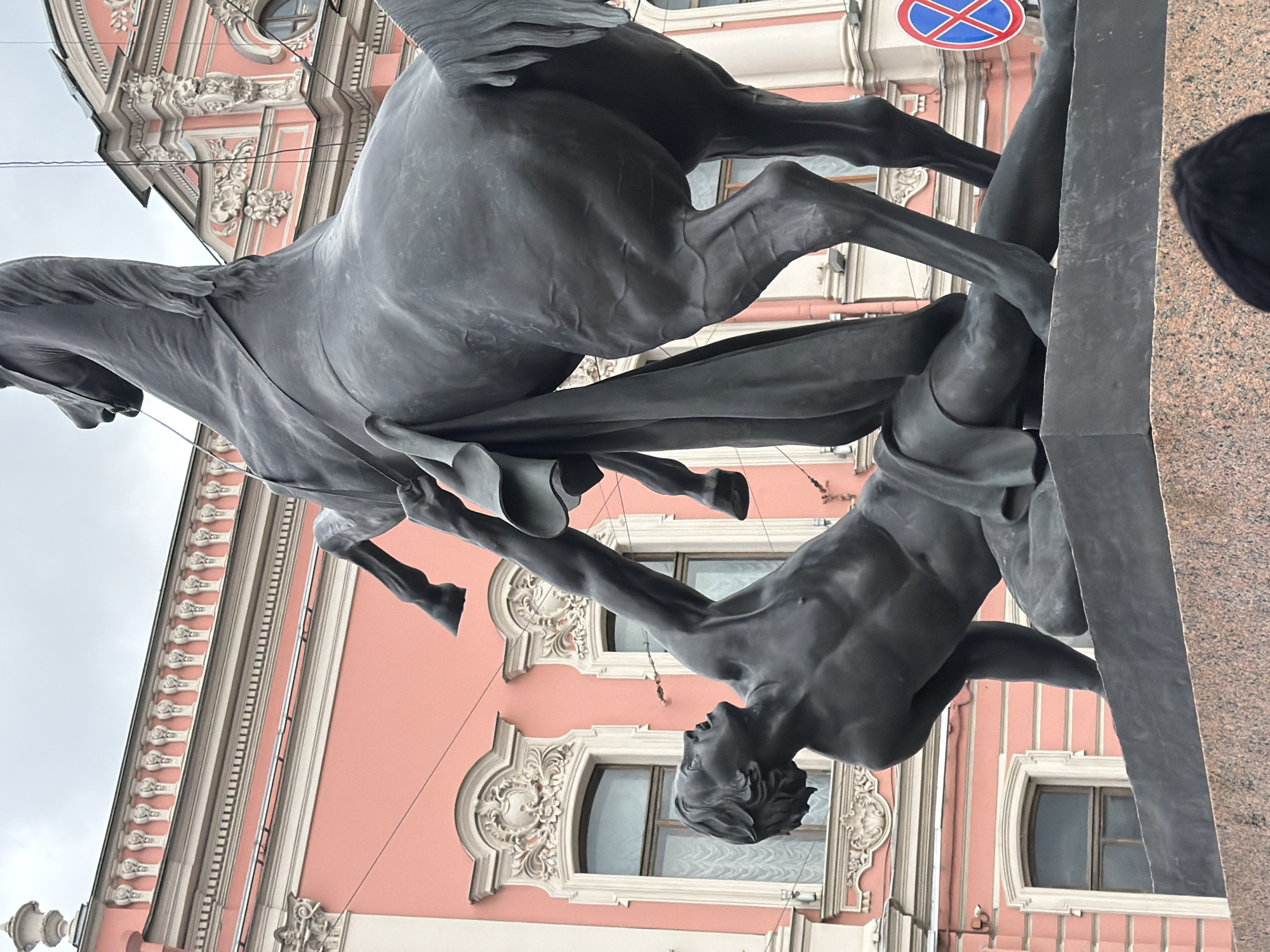 The media file depicts a dramatic bronze statue of a man and a winged horse, likely Pegasus, set against the backdrop of an ornate building. The scene captures a dynamic struggle, with the statue positioned such that the horse rears high, its powerful form dominating the composition, and the man grappling beneath it.
The central subjects are a powerful, muscular man and a rearing horse. The man is depicted nude, seemingly in a struggle, positioned beneath the horse with his head thrown back and mouth open in an expression of exertion or agony. His arms are extended towards the horse. The horse, identifiable by its wings and majestic pose, is rearing dramatically on its hind legs, its front hooves lifted high, showcasing its powerful musculature and flowing mane and tail. The scene captures an intense moment, possibly Bellerophon grappling with Pegasus.
In the background, a grand, multi-story building with a salmon-pink facade dominates. It is adorned with rich white classical architectural details, including elaborate window frames, cornices, pilasters, and decorative carvings like festoons and rosettes. Above the building, a grey, overcast sky is visible, suggesting daytime. A red and blue circular road sign, likely a 'no entry' or 'prohibited' sign, is visible in the upper right background, adjacent to the statue. A mottled brown and pink granite base supports the statue, parts of which are visible on the right. A dark-haired person's head is partially cropped into the lower right corner, indicating other people present at the scene. The overall impression is one of dynamic struggle captured in art, set within a historic urban environment like Saint Petersburg, Russia.