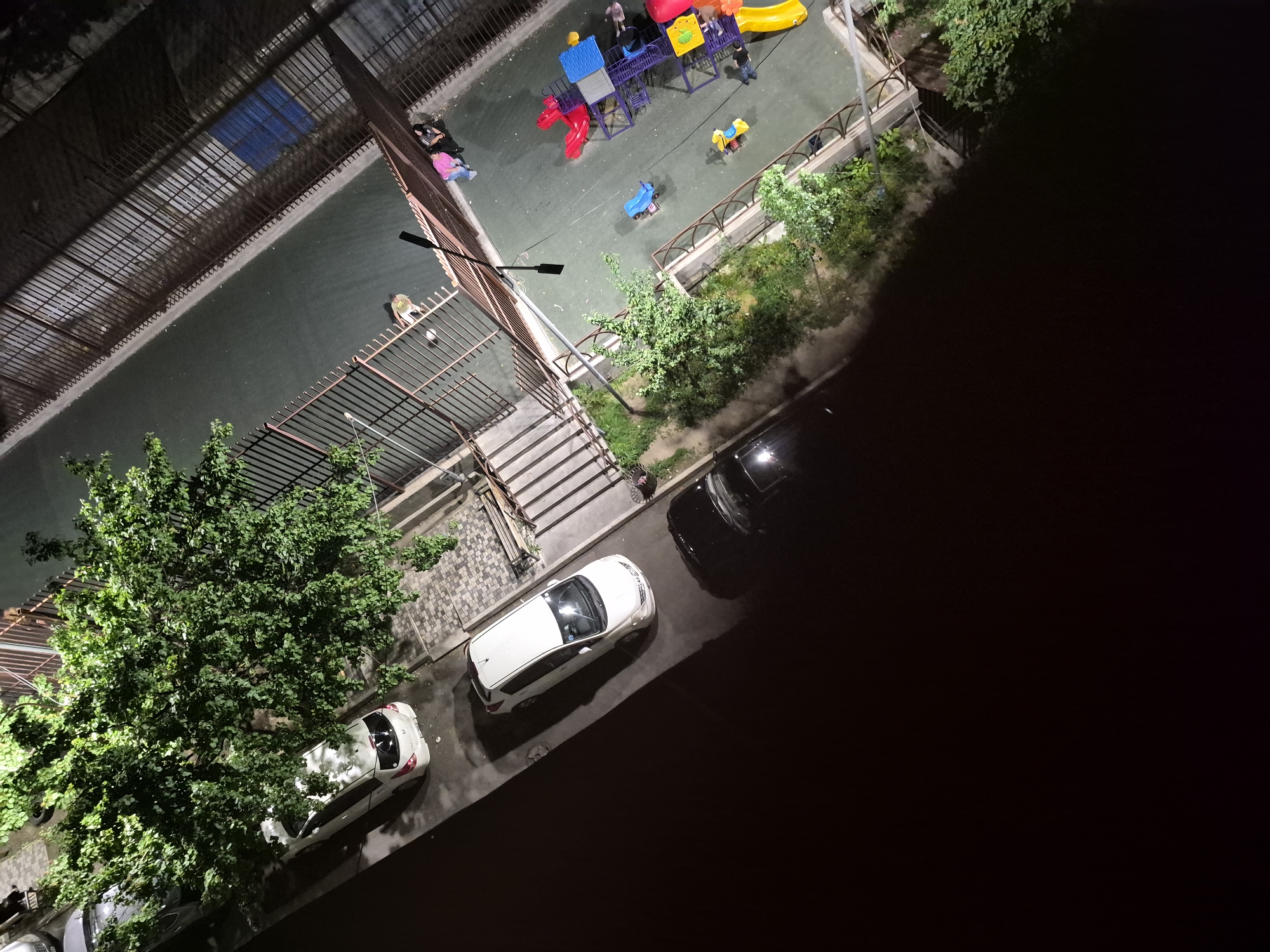 This elevated night shot depicts a vibrant playground and adjacent areas in a residential setting in Yerevan, Armenia. The scene is illuminated by artificial lights, casting shadows and highlighting specific features.
In the center, a colorful children's playground features various equipment, including red and yellow slides, a blue and purple climbing frame, and a blue scooter and yellow animal-shaped spring riders. Several individuals are present: two people, one in a pink top, are seated near a fence at the playground's edge. Further in, adults and at least one child are near the play structures. Adjacent to the playground, a fenced-off green area or sports court hosts a person in light clothing sitting on the ground near a small white ball.
The foreground and left side of the image show a street with multiple parked cars. A white SUV or minivan is prominently parked, with parts of other white, dark, and silver vehicles visible. A large, leafy green tree overhangs the street and partially obscures one car. Smaller trees and bushes line the fences surrounding the playground and green area. A street lamp extends into the frame, providing illumination. Concrete steps with paving stones lead down from an unseen elevated area to the street level. The image conveys a calm, everyday urban scene after dark. No visible text is clearly discernible.