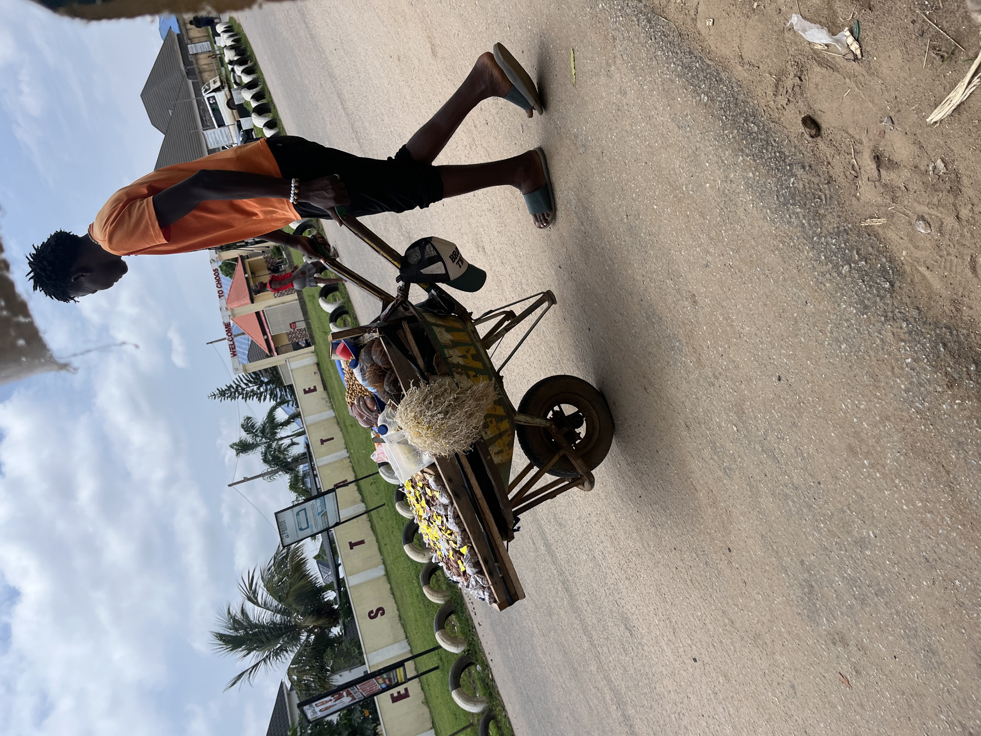 A young Black male wearing an orange t-shirt and black pants pushes a wooden cart filled with produce and packaged goods along a paved road. He is wearing dark-colored slides on his feet and a beaded bracelet on his left wrist. A baseball cap hangs from the cart's handle. The cart is laden with various items, including what appear to be fruits like coconuts, small packaged goods, and a bundle of dried grass or straw. In the background, a building with a "WELCOME TO CHOOS" sign is visible, along with a white van and other structures. Several palm trees and tire planters line the roadside. The scene appears to be in Abuja, Nigeria, given the specific details in the signage and vegetation.