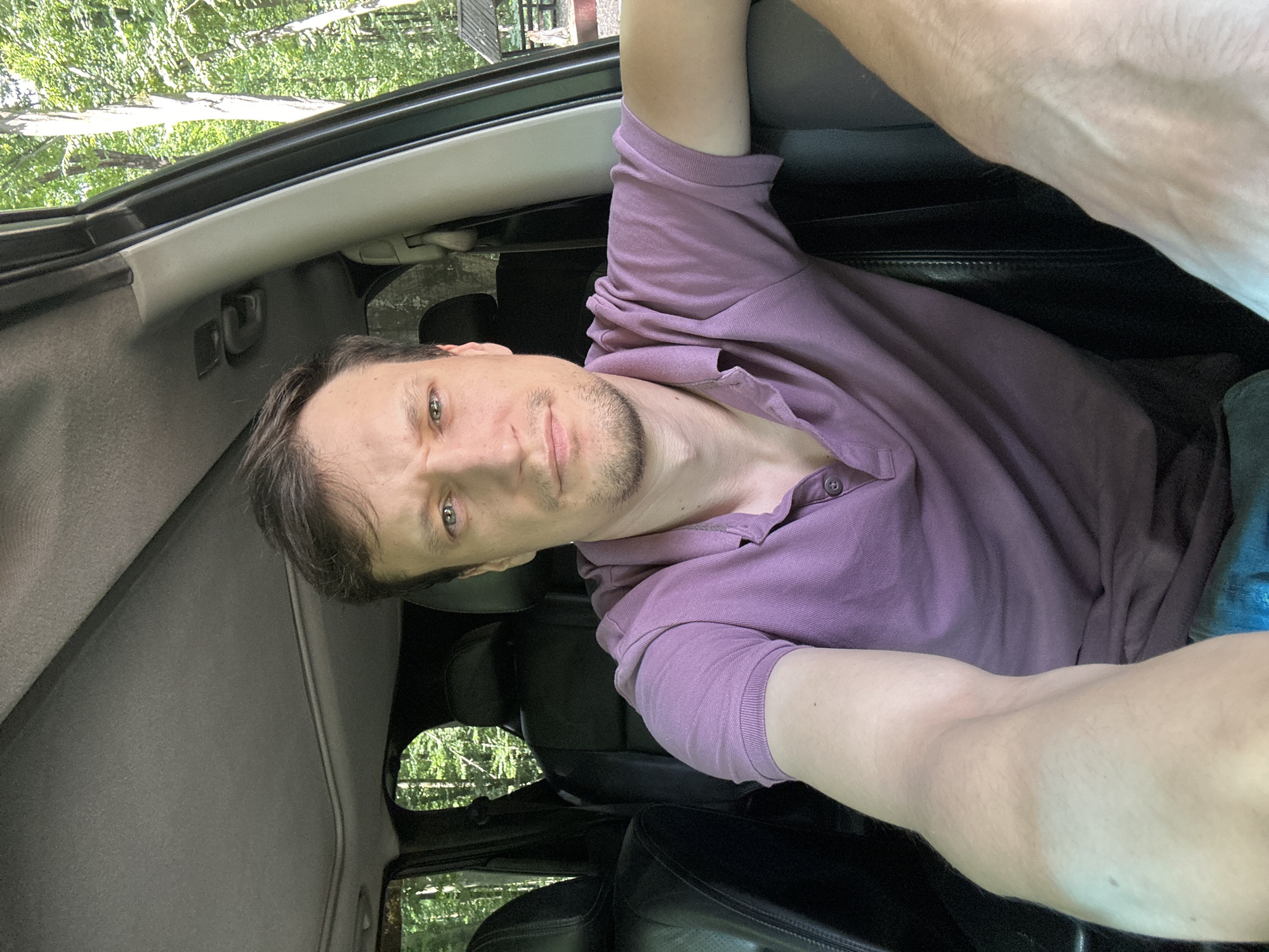 In Shatava, Ukraine, a man is taking a selfie from inside a car. He has short brown hair and is wearing a purple polo shirt. The car's interior is visible, with black leather seats and a sunroof. The background shows a wooded area with green trees and a wooden structure in the distance.