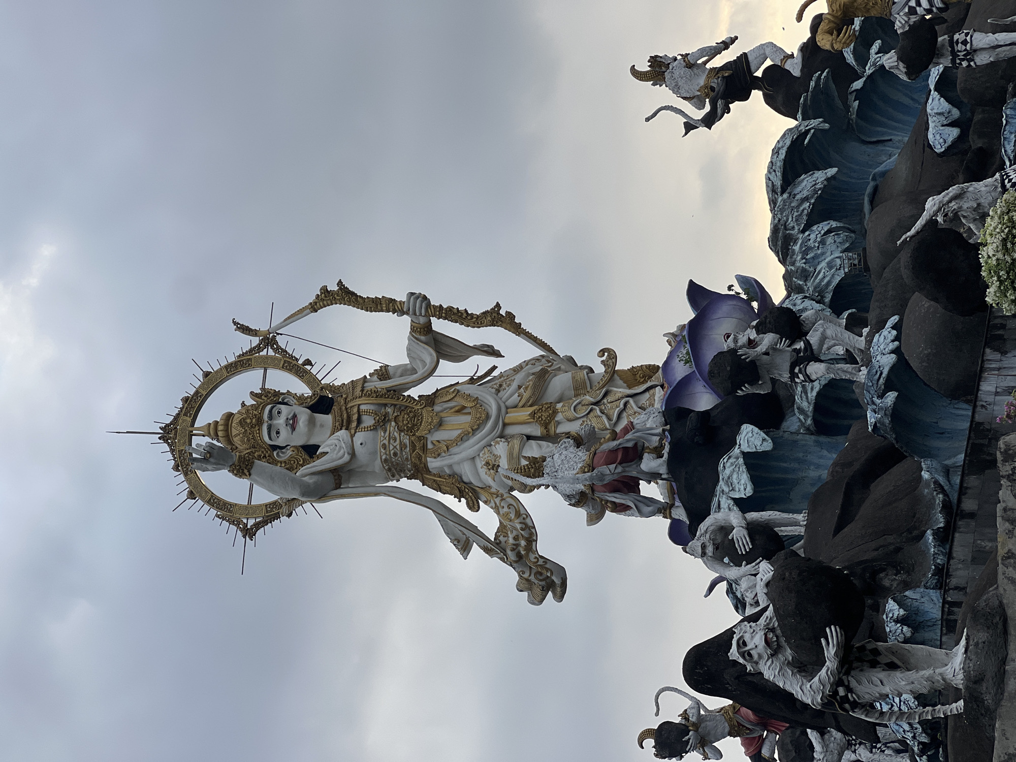 This is a low-angle, wide shot of a large, ornate statue in Denpasar, Indonesia, under a cloudy sky. The main subject is a towering, white and gold statue of a deity holding a bow and arrow, with a golden halo behind its head. The deity wears elaborate clothing and jewelry. Below the main statue, a smaller figure in a red and white outfit kneels on a dark, lotus-like base. Surrounding this central structure is a collection of other statues, many of them appearing to be monkey-like figures made of white stone. Some of these figures are depicted in various poses, some carrying dark, rounded objects resembling rocks. The entire monument is set against a backdrop of dark, wave-like rock formations with accents of blue. In the background, on the right side of the frame, another white and gold statue of a figure in a dynamic pose is visible. The overall scene is grand and appears to be an outdoor religious or cultural monument.