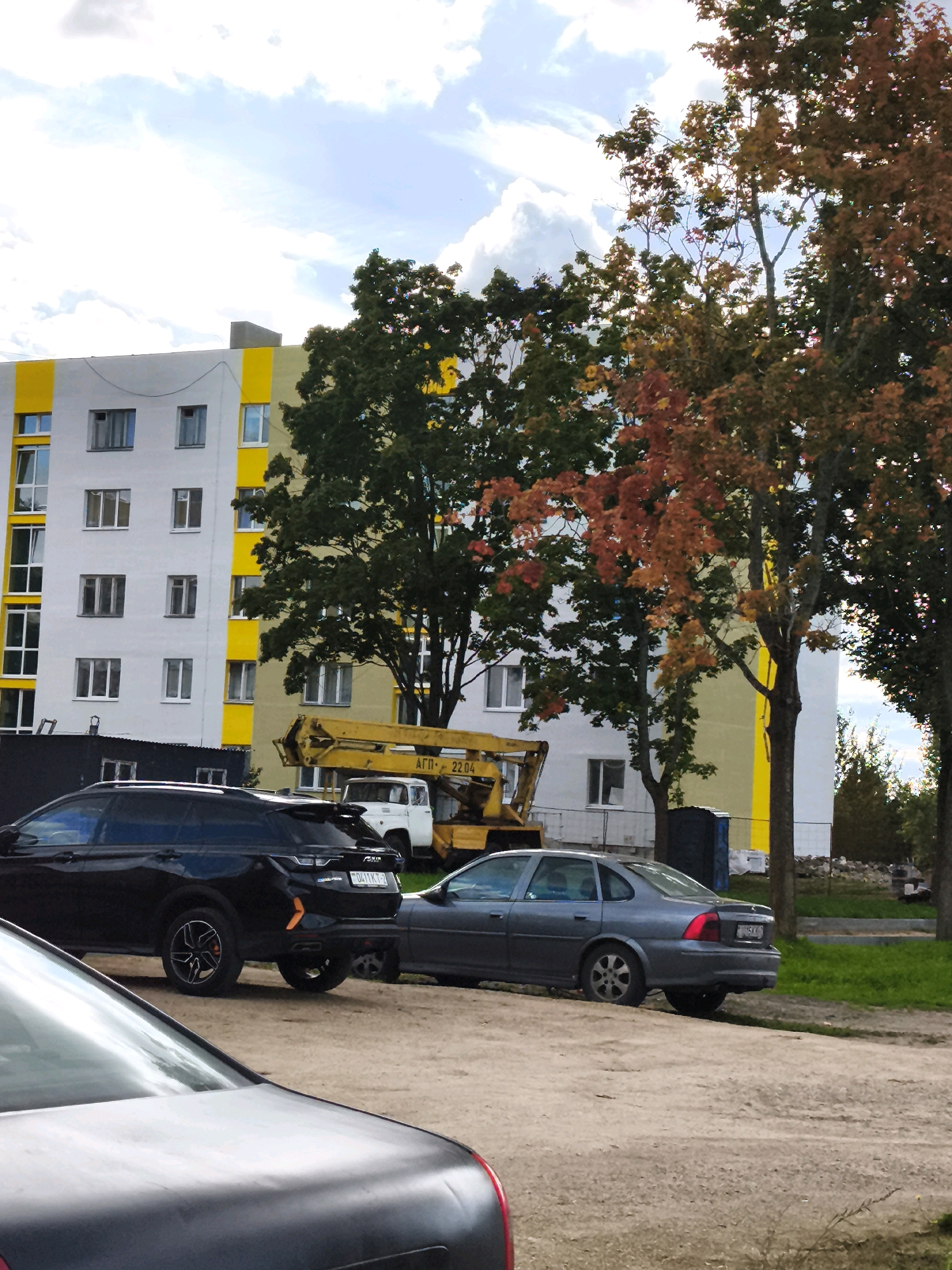 The image shows a residential building with yellow and white accents, characteristic of Soviet-era architecture, possibly located in a rural or suburban area of Горскі сельскі Савет, Belarus. In front of the building, several vehicles are parked on a dirt road. These include a black SUV, a grey sedan, and a white utility vehicle. A yellow construction crane is also visible, suggesting ongoing work or recent construction activity. The scene is framed by trees with some autumn foliage, indicating the season. The sky is partly cloudy.