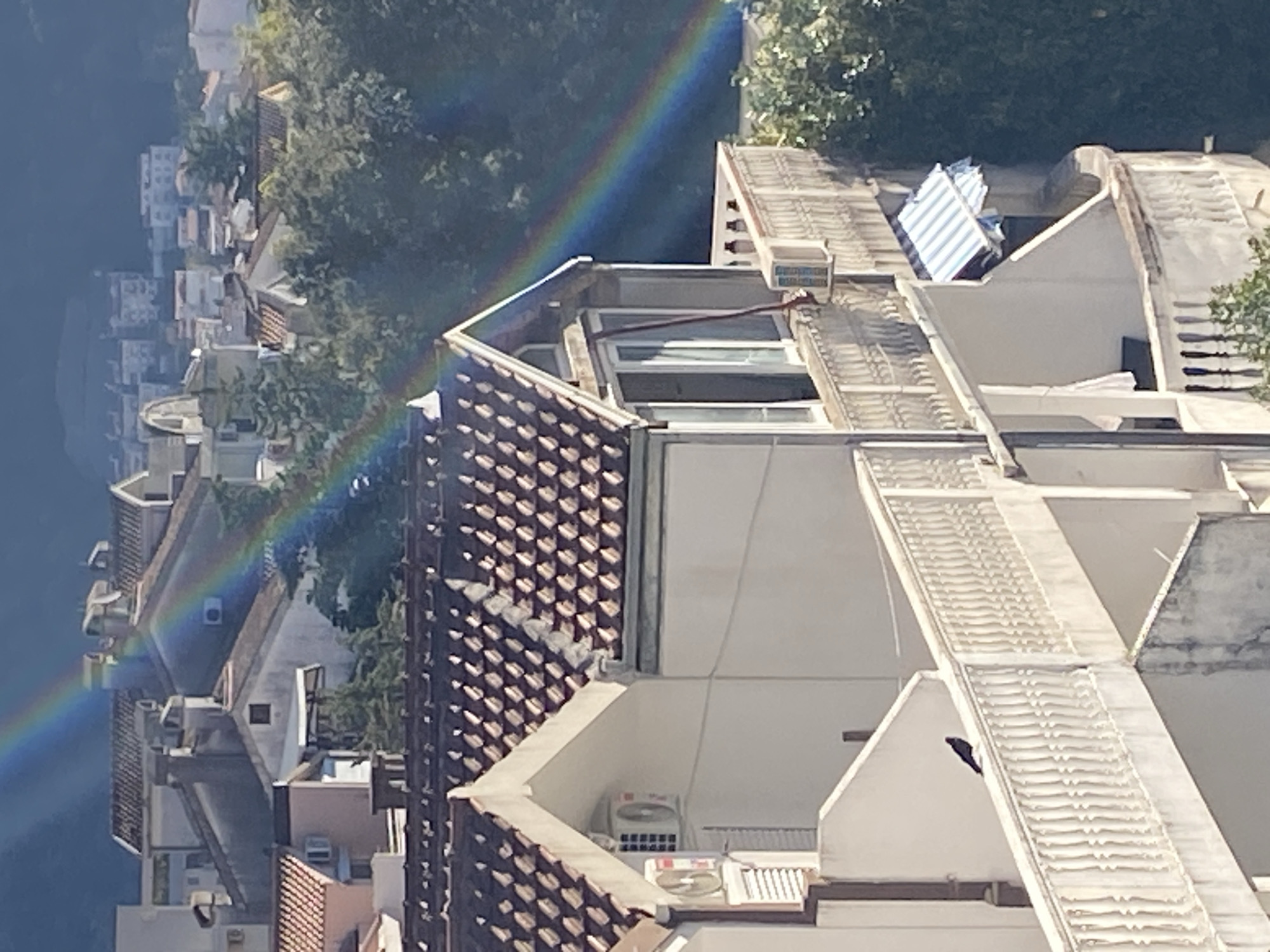 The image depicts an aerial view of a section of Budva, Montenegro, characterized by densely packed buildings with a mix of white-walled structures and tiled roofs. In the foreground are several multi-story buildings, exhibiting air conditioning units and window openings. A portion of a rainbow is visible in the upper left corner of the image, extending downwards across the urban landscape. The overall environment suggests a sunny day with good visibility, and the scene shows the typical architecture and layout of a Mediterranean town. The angle of the shot and close proximity to the buildings obscure most street-level activity.