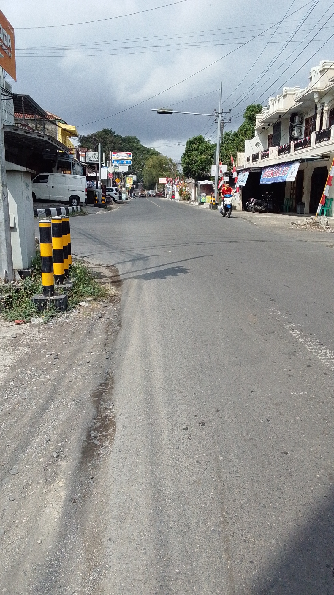 The image depicts a street scene in Desa Karangbenda, Indonesia. On the left side of the road, there is a white van parked next to a building with an awning. Several black and yellow bollards are positioned along the side of the road. Further down the road, there are buildings with shops and signs, including one that appears to be a convenience store with an Indomaret logo. A person is riding a scooter with a red shirt down the middle of the road. The road itself is paved and appears to be a two-lane street with some patches of gravel on the shoulder. The sky is overcast with some clouds. Trees are visible in the background. The architecture of the buildings suggests a tropical or sub-tropical climate.