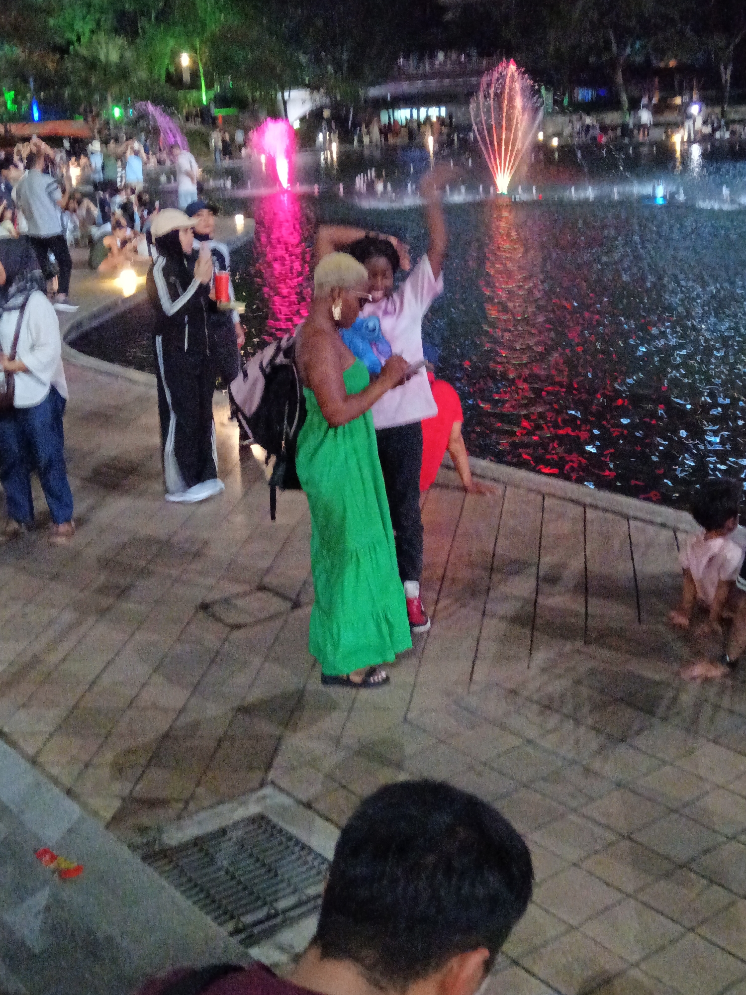 The image depicts a vibrant night scene in Kuala Lumpur, Malaysia, near the iconic Petronas Twin Towers. A group of people are gathered around a fountain that illuminates the dark water with pink and red hues. The foreground features a wooden walkway where several individuals are present.
In the center, a woman with blonde hair, wearing a bright green strappy dress, is holding a blue object. Next to her, another person, partially visible, is wearing a pink t-shirt. To the left of these two, a person in a black tracksuit with white stripes and a beige headscarf is holding a red cup. Further to the left, a person in a white top and blue pants is holding a bag. In the background, numerous other people are milling around, some seated, and the silhouette of buildings and trees can be seen. The atmosphere is lively, suggesting a public gathering or event.