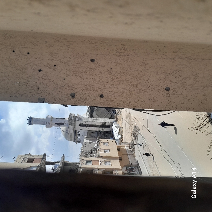 The image presents a street scene in Khan Yunis, Palestinian Territory, marked by extensive destruction. In the background, the Great Mosque of Khan Yunis stands prominently, its white dome and minaret showing significant structural damage, with sections of the dome and upper minaret compromised. Adjacent to the mosque, and extending along the left side of the street, are multiple multi-story buildings, severely damaged, with large portions of their facades missing, exposing interiors and twisted rebar. The paved street below is clear of large debris but appears dusty, with several electrical or communication wires hanging low across it. On the immediate right, a light beige, textured wall, likely part of an adjacent building, displays multiple distinct circular indentations or impact marks. Three individuals are visible on the street: one person in dark clothing walks in the mid-ground, moving away from the viewer, while two other figures are further down the street on the left, also walking away. The sky above is partly cloudy.