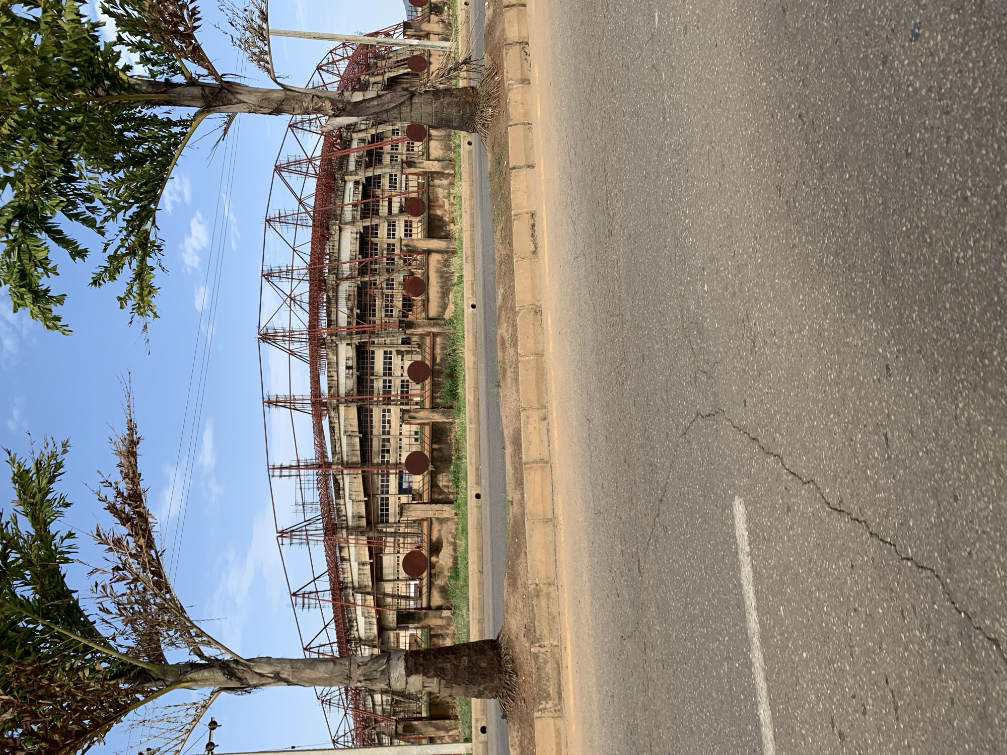 The image shows a section of road in Rafin Par, Nigeria. The road has a white line marking and several cracks. On the left and right sides of the road are sidewalks lined with palm trees. Beyond the sidewalks is a large building under construction with scaffolding visible. The sky is blue with some clouds.