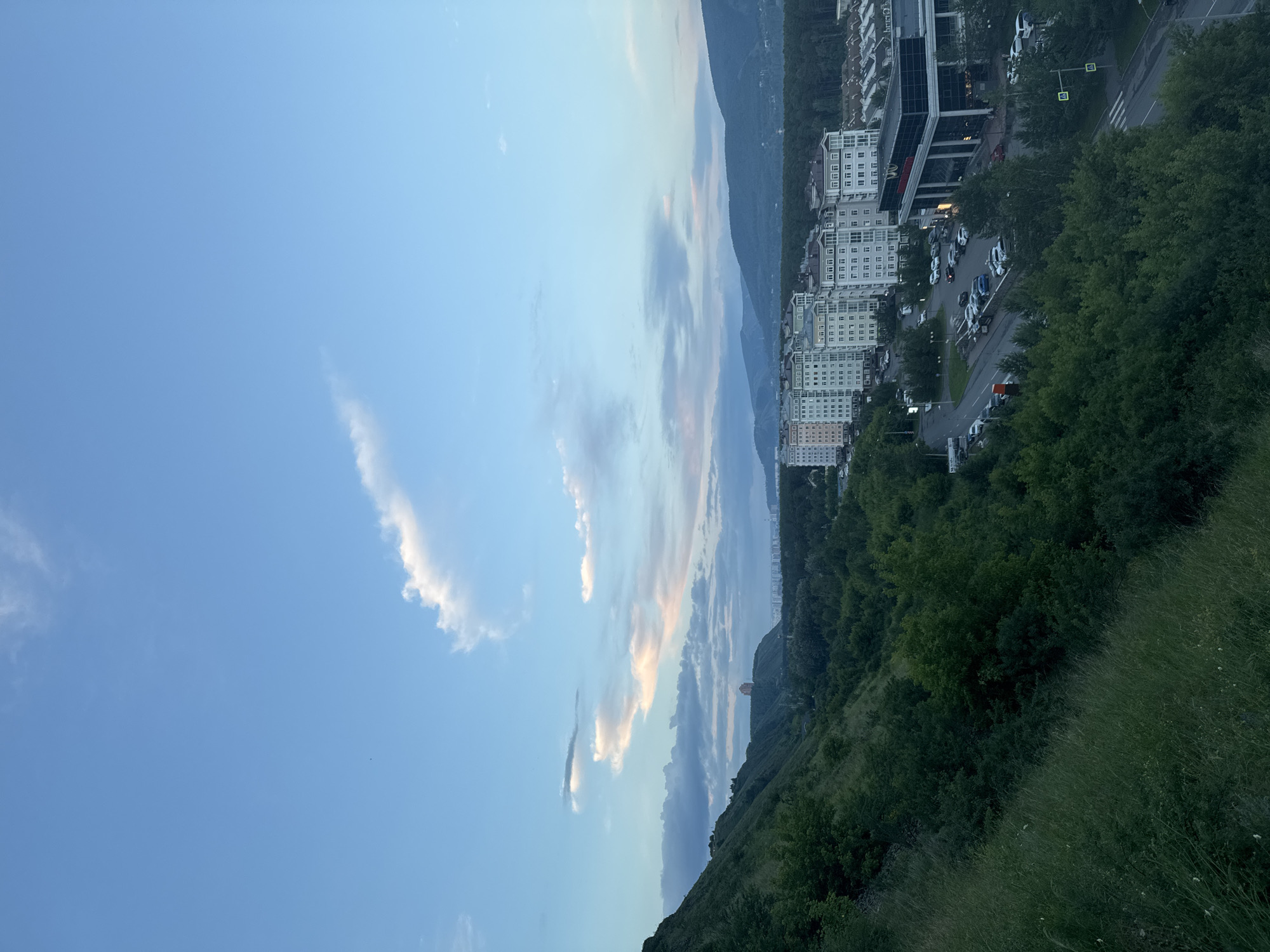 This is a vertical, eye-level, outdoor, daytime photograph of a city nestled at the base of a mountain range, with a clear blue sky and scattered clouds above. The city features numerous multi-story buildings, some with white facades and numerous windows, characteristic of apartment complexes. A paved road with cars parked on the side and driving on it winds through the urban landscape. Tall, dense green trees line the road and extend up the slopes of the mountains, creating a strong contrast between the built environment and the natural landscape. The mountain slopes are covered in lush vegetation, and the sky shows hints of sunset colors, suggesting the time is late afternoon or early evening. The overall atmosphere is peaceful and serene, with a hint of urban life integrated into a natural setting. There is no visible text in the image.
